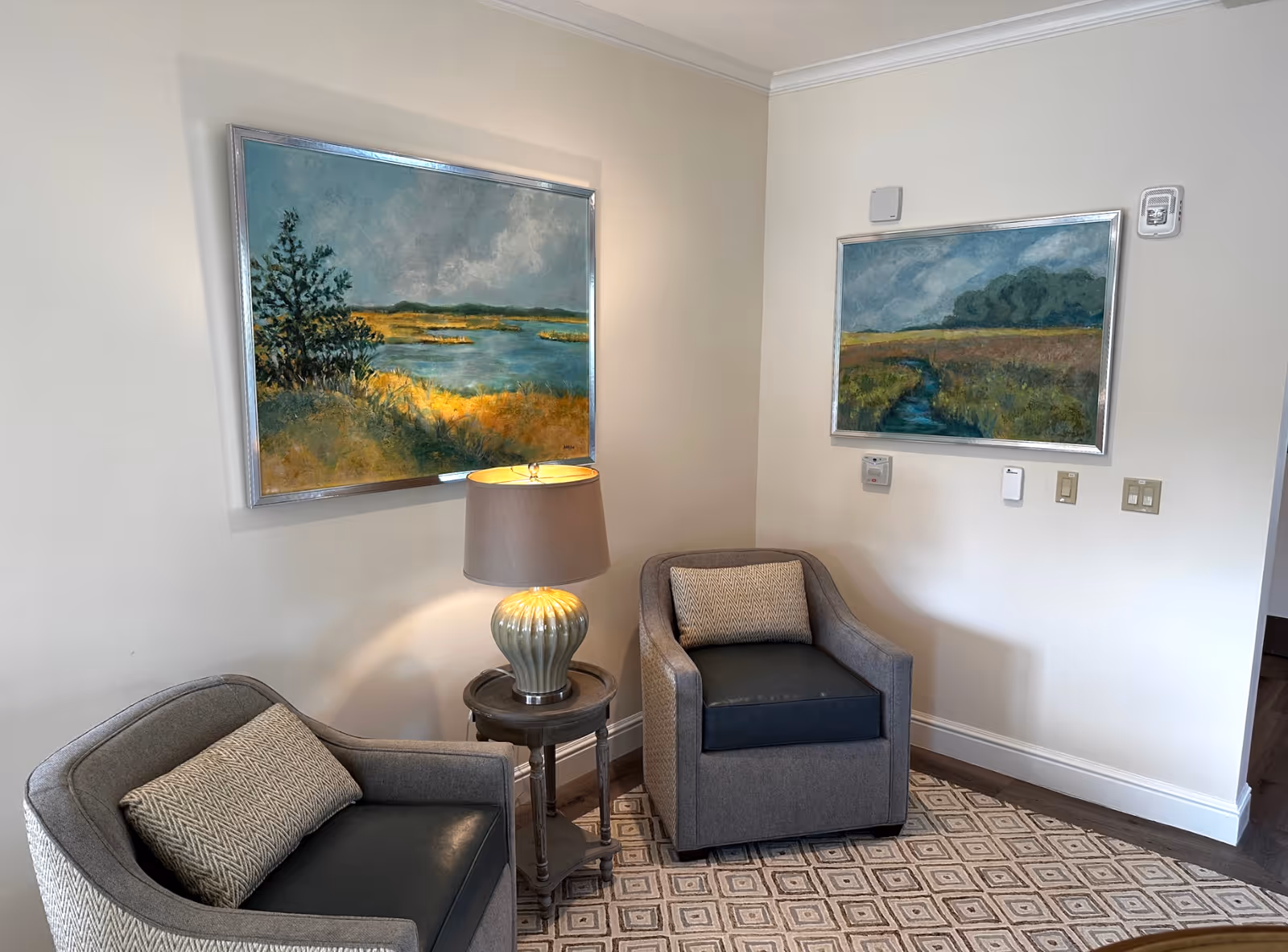 A cozy corner of a room with two gray upholstered armchairs, each with a patterned cushion. Between the chairs is a small round wooden side table with a ceramic lamp that is turned on. The walls are light-colored and adorned with two framed landscape paintings depicting nature scenes with water and greenery. The floor has a patterned area rug.