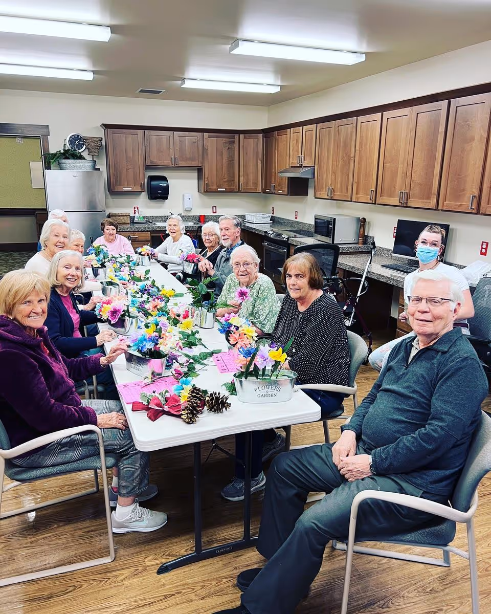 A group of elderly people sitting around a long table in a kitchen area, engaging in a flower arranging activity with colorful artificial flowers and pine cones. A staff member wearing a mask is standing in the background near the counter. The room has wooden cabinets, a refrigerator, microwave, and bright overhead lighting.