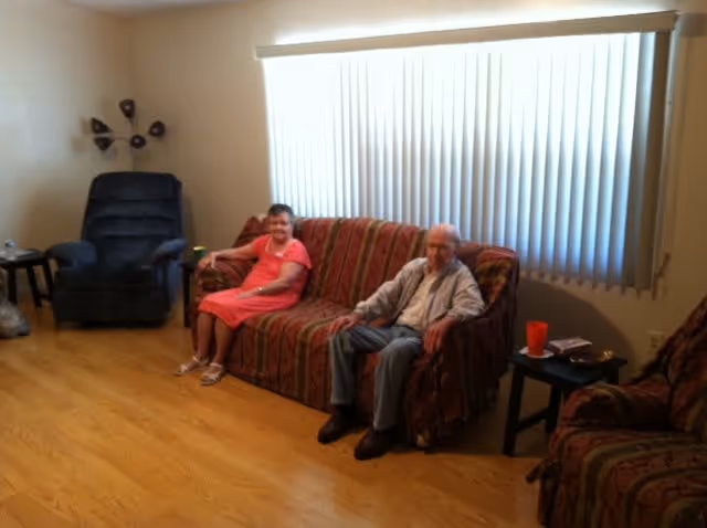 An elderly man and woman sitting on a patterned sofa in a living room with wooden flooring. The room has a large window with vertical blinds, a black recliner chair, a side table with a red cup, and a wall-mounted decorative piece.