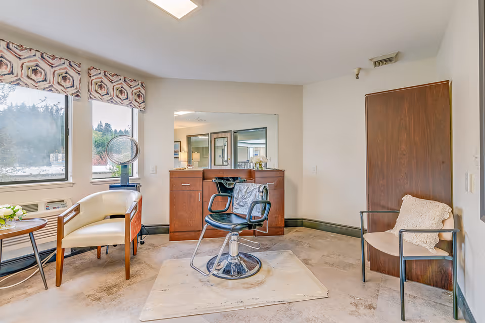 A small salon area inside a senior living facility with a black salon chair on a protective mat in front of a wooden vanity with a large mirror. There are two chairs on either side of the room, one white with wooden arms and the other beige with a crocheted pillow. Two windows with patterned valances let in natural light, and a hair dryer is positioned near one window.