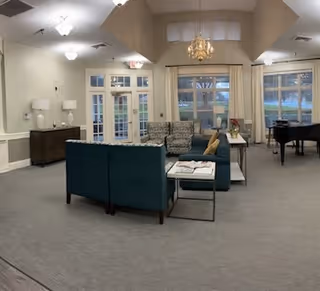 Spacious senior living common room with sofas and chairs arranged around tables, a piano, chandelier, and large windows.