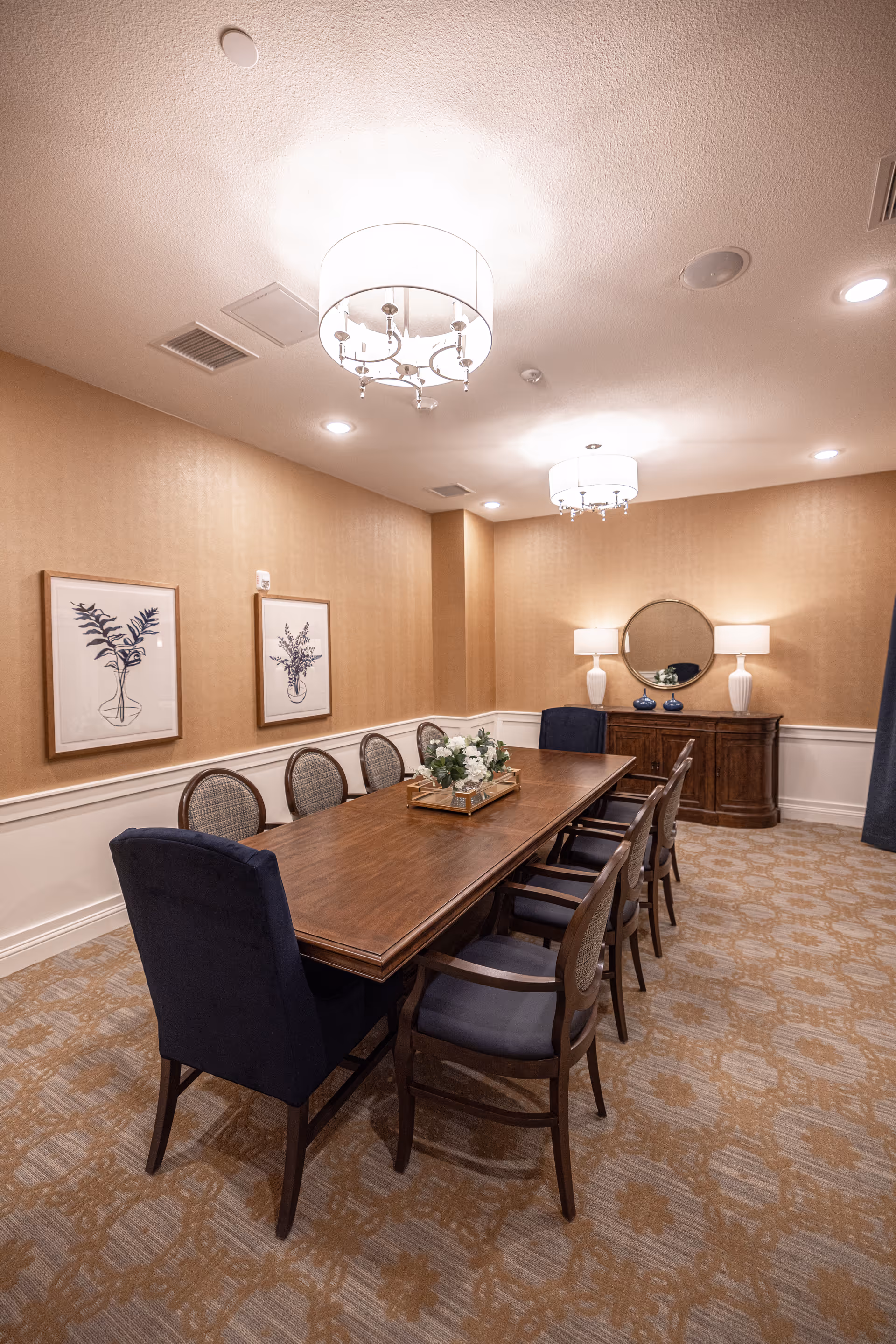 A well-lit dining room with a long wooden table surrounded by eight chairs, two of which have dark blue upholstery. The room features beige walls with white wainscoting, two framed botanical prints on one wall, a wooden sideboard with a round mirror above it, and two white table lamps. The carpet has a patterned design in neutral tones, and two modern ceiling light fixtures hang above the table.