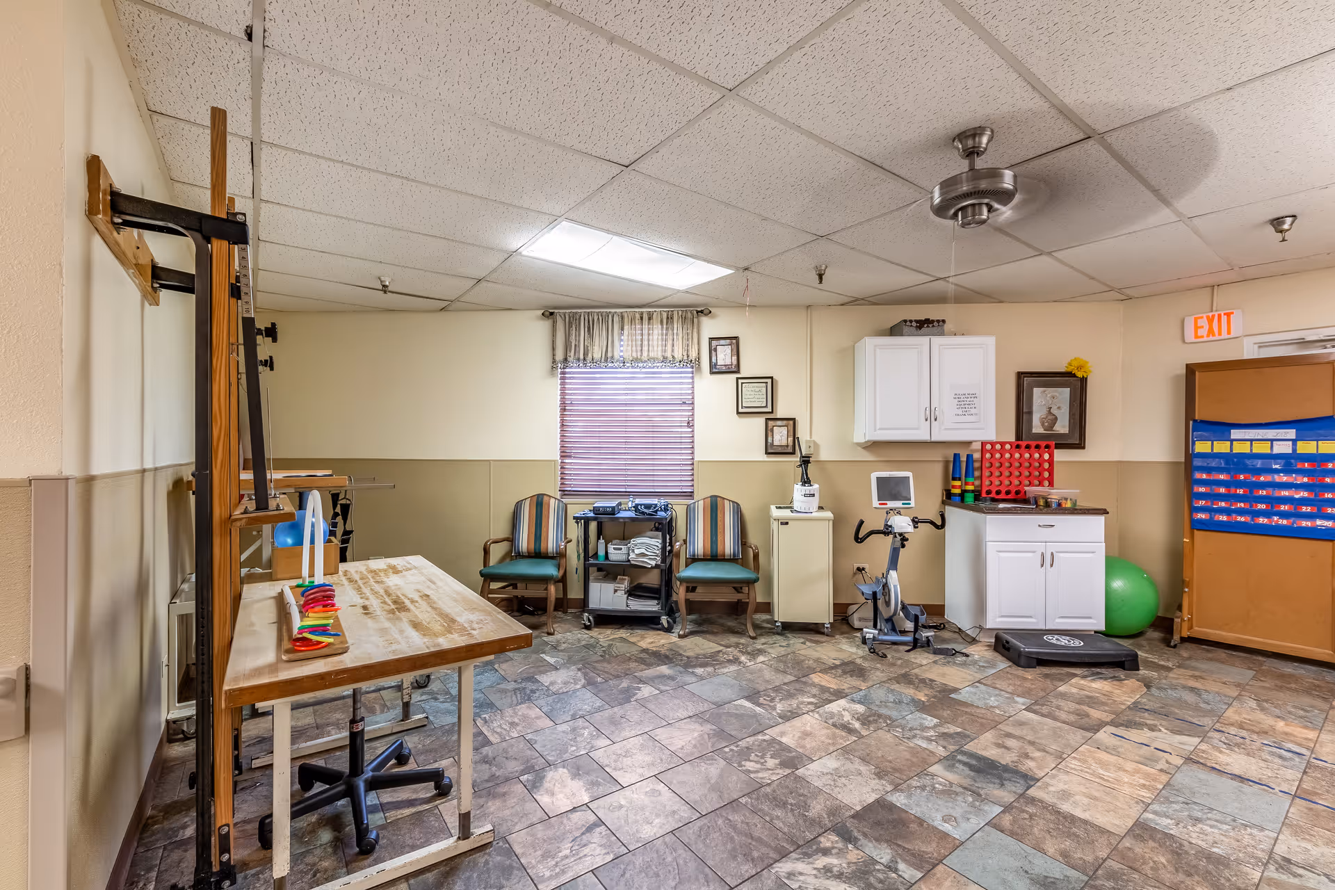 A nursing home activity/therapy room with chairs, exercise equipment, a table, cabinets, and a window.