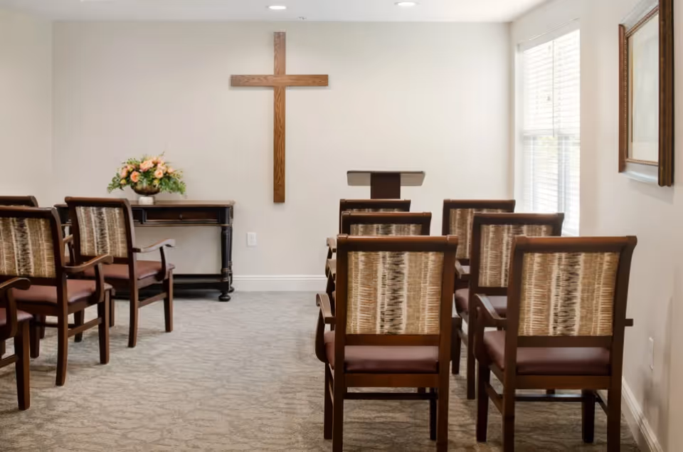 Small chapel-style room with rows of wooden chairs facing a lectern and a wall-mounted wooden cross.