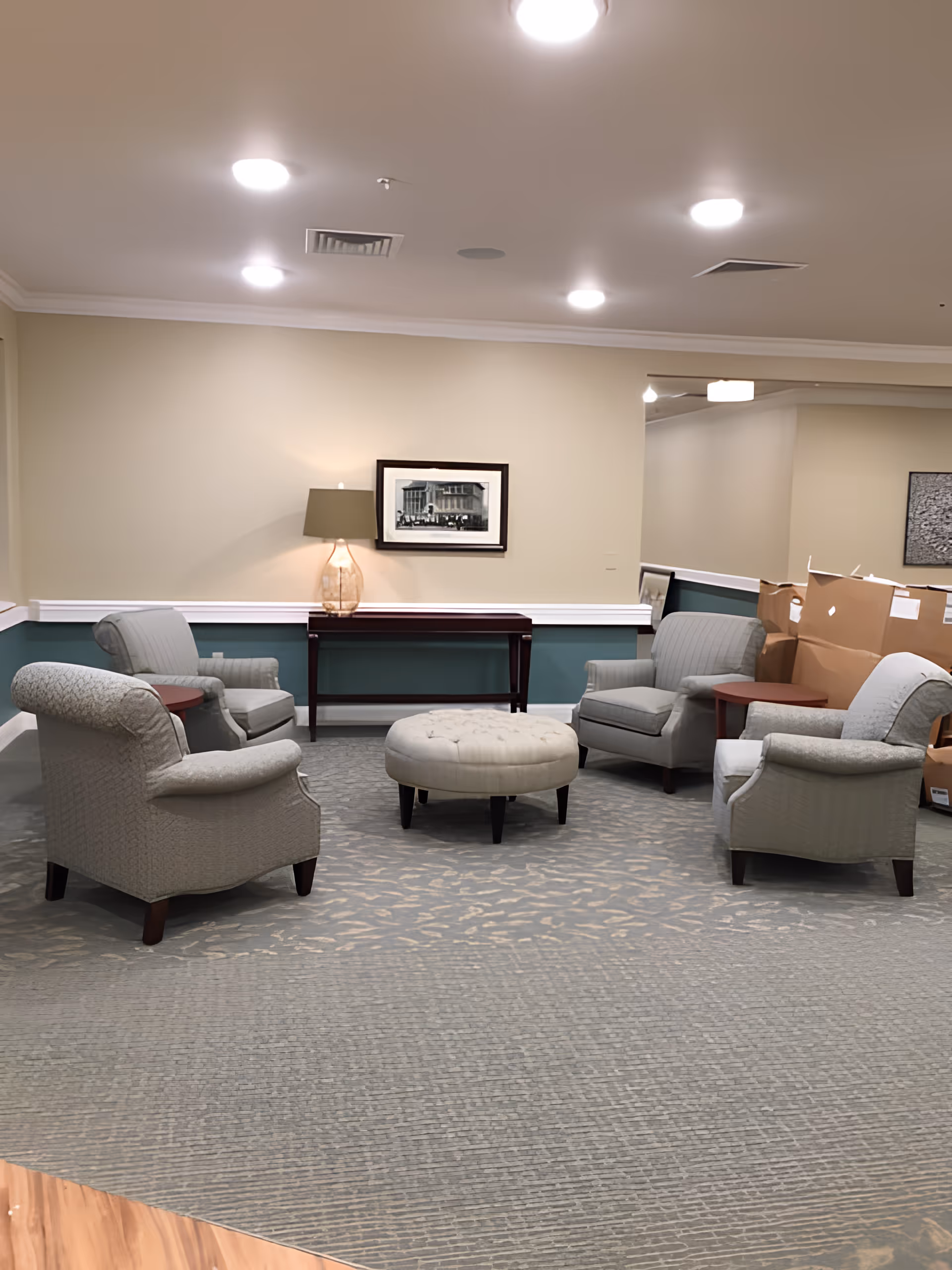 A lounge area with four upholstered armchairs arranged around a round ottoman, a console table with a lamp and framed picture against a beige wall.