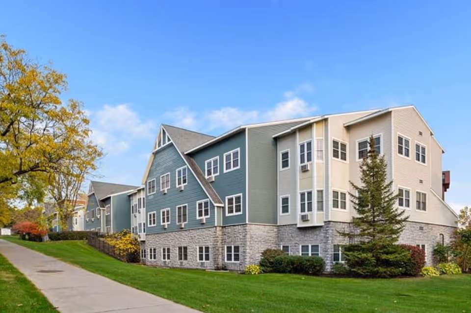 Exterior view of a multi-story senior living facility building with green and beige siding, stone foundation, multiple windows, and a well-maintained lawn with trees and shrubs under a blue sky.