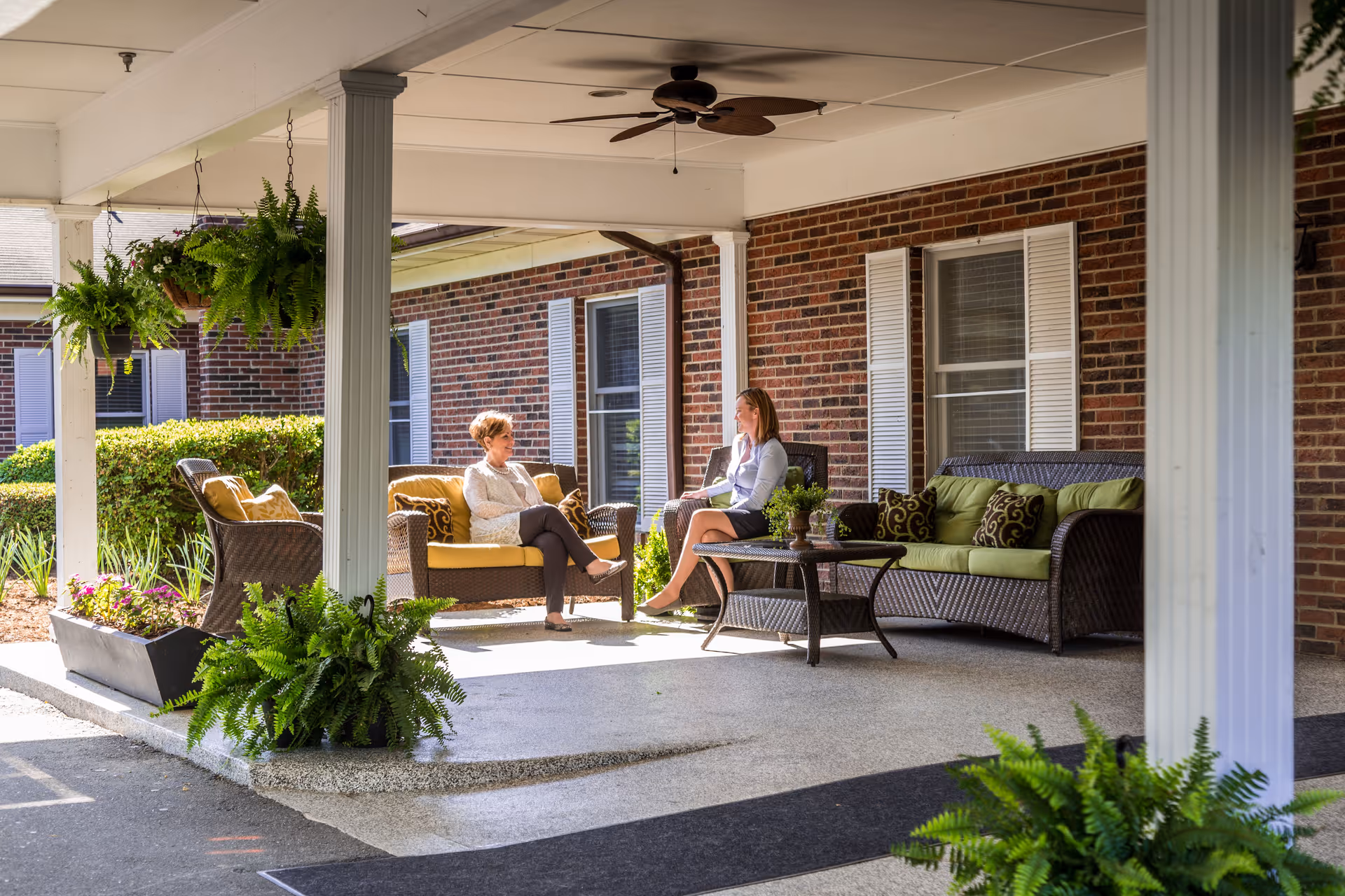 Two women sit and talk on wicker seating under a covered brick entry porch with potted ferns and hanging plants.