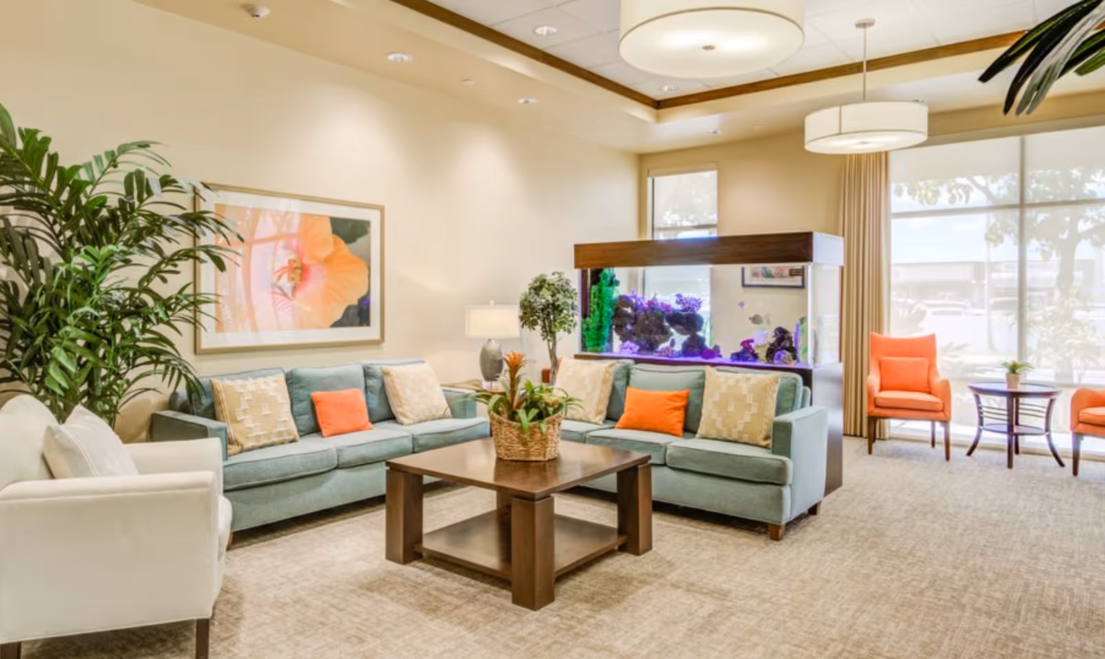 A bright and spacious living room with two light blue sofas adorned with beige and orange pillows, a white armchair, a wooden coffee table with a plant centerpiece, a large aquarium with colorful coral and fish, two orange chairs with a small round table between them near large windows, and decorative plants and artwork on the walls.