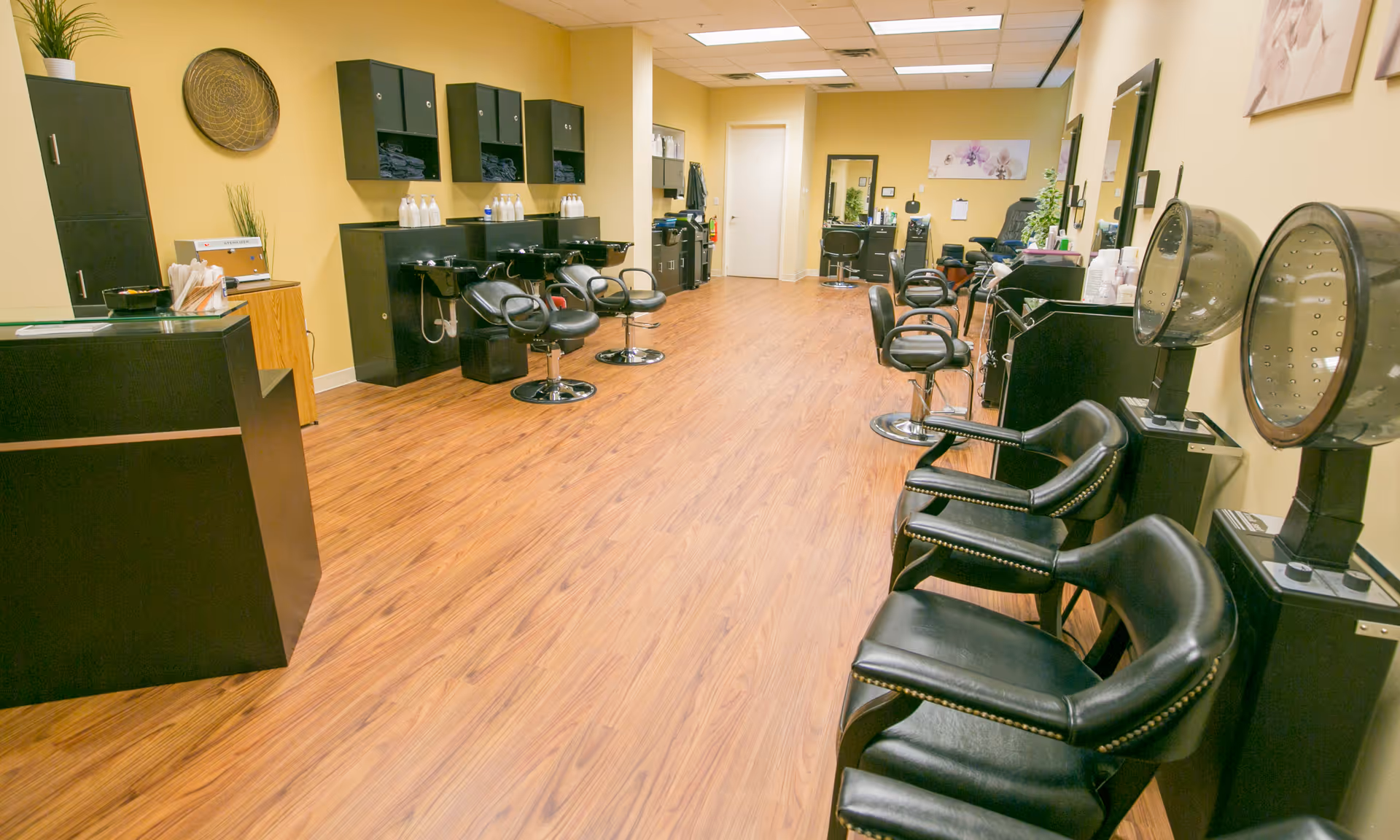 Interior view of a hair salon area in a healthcare facility with black salon chairs, hair washing stations, hair dryers, and wooden flooring. The walls are painted yellow and decorated with plants and artwork.