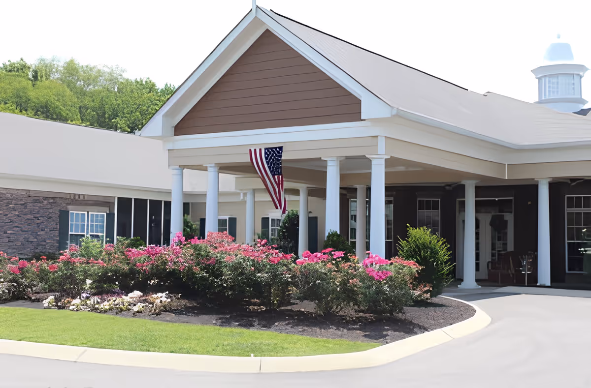 Entrance of a senior living facility with a covered driveway supported by white columns, an American flag hanging from the ceiling, and a landscaped garden with pink flowers and green shrubs in front.