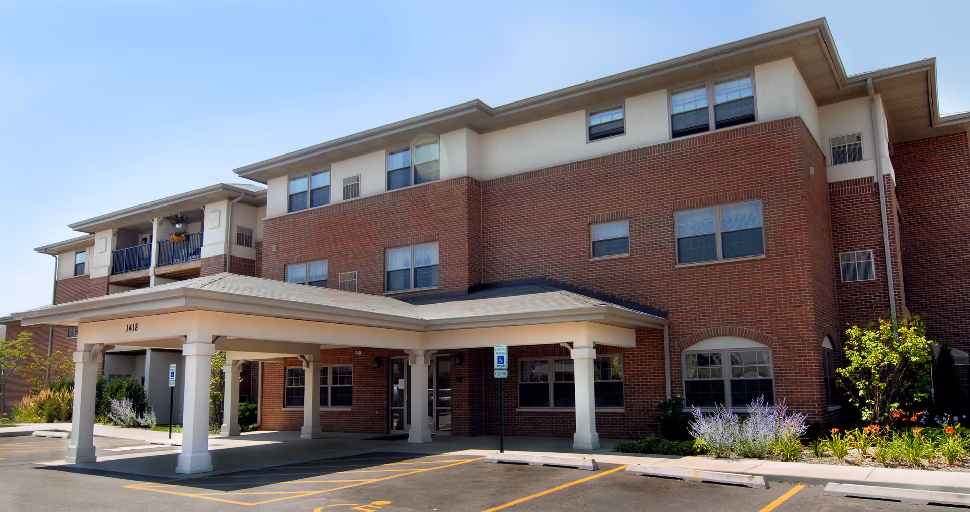 Exterior view of a three-story brick building with a covered entrance and several windows. There are parking spaces in front, including a handicapped parking spot, and some landscaping with bushes and flowers near the building.