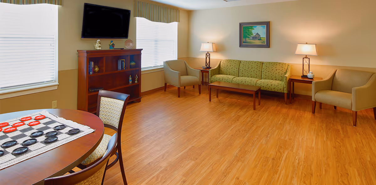 Bright common sitting room with a green sofa, armchairs, a TV on a wooden cabinet, a round game table with checkers, and wood flooring.