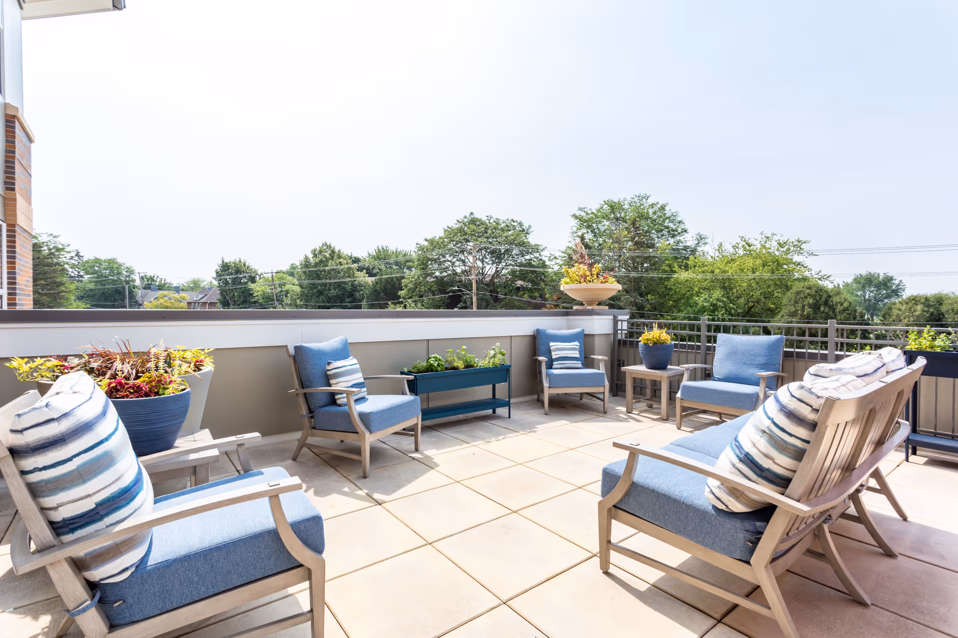 Outdoor patio area with cushioned wooden chairs and a small table, decorated with potted plants and flowers, overlooking trees and a clear sky.