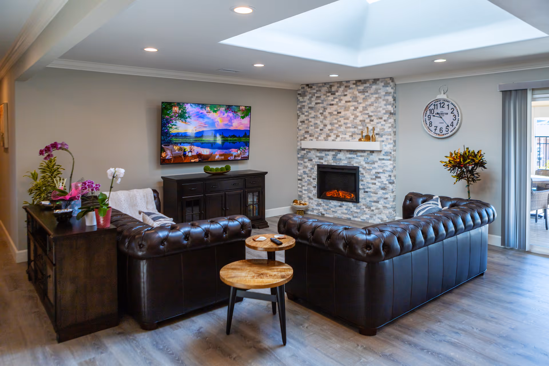 A cozy living room with two dark brown leather tufted sofas arranged around a small round wooden coffee table. A modern electric fireplace with a stone tile surround is on the wall, above which is a white mantel with decorative items. A flat-screen TV is mounted on the adjacent wall, displaying a colorful nature scene. There is a large wall clock and a plant in the corner near sliding glass doors that lead to an outdoor area. The room has recessed lighting and light wood flooring.