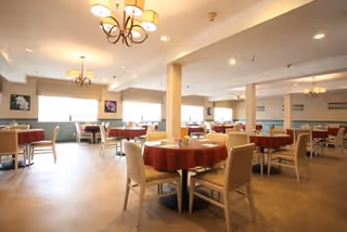 A spacious dining room with multiple round tables covered with red tablecloths and surrounded by beige chairs. The room is well-lit with ceiling lights and chandeliers, and large windows allow natural light to enter. The walls are decorated with framed artwork.