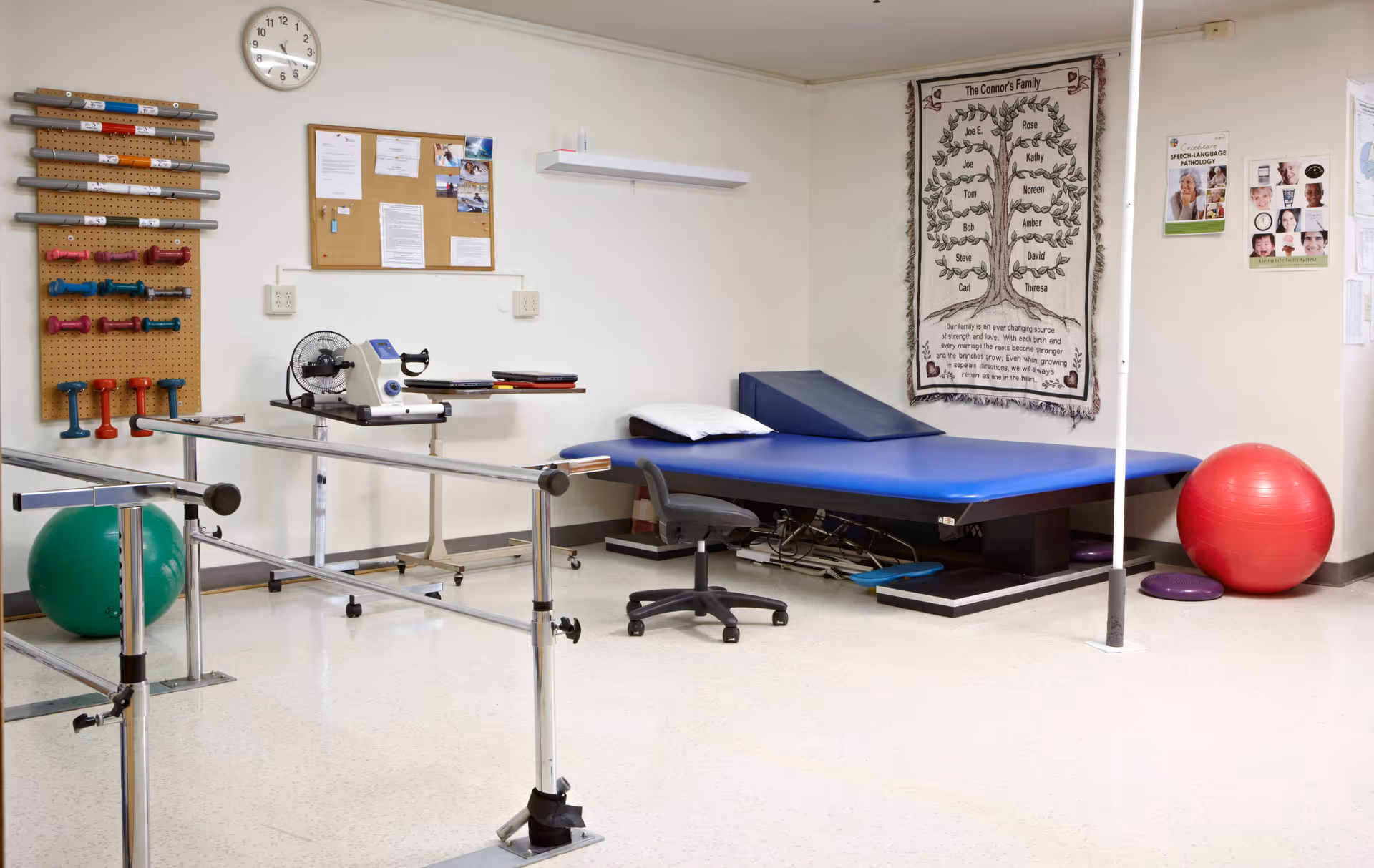 Physical therapy room with parallel bars for walking exercises, a blue therapy table with a pillow and wedge, a black rolling chair, various exercise equipment including dumbbells and exercise balls, a clock on the wall, a bulletin board, and posters on the walls.