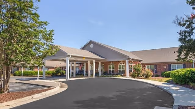 Front exterior view of a single-story brick building with a covered entrance and a circular driveway, surrounded by trees and shrubs under a clear blue sky.