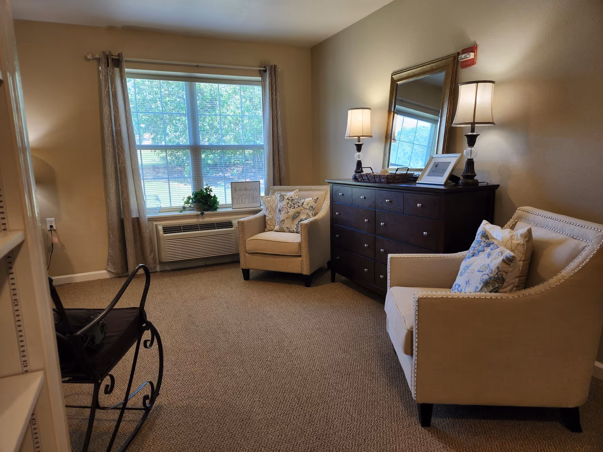 A cozy sitting area with two beige armchairs adorned with patterned cushions, a dark wooden dresser with two table lamps and a large mirror above it, a window with curtains letting in natural light, and a rocking chair partially visible on the left side.