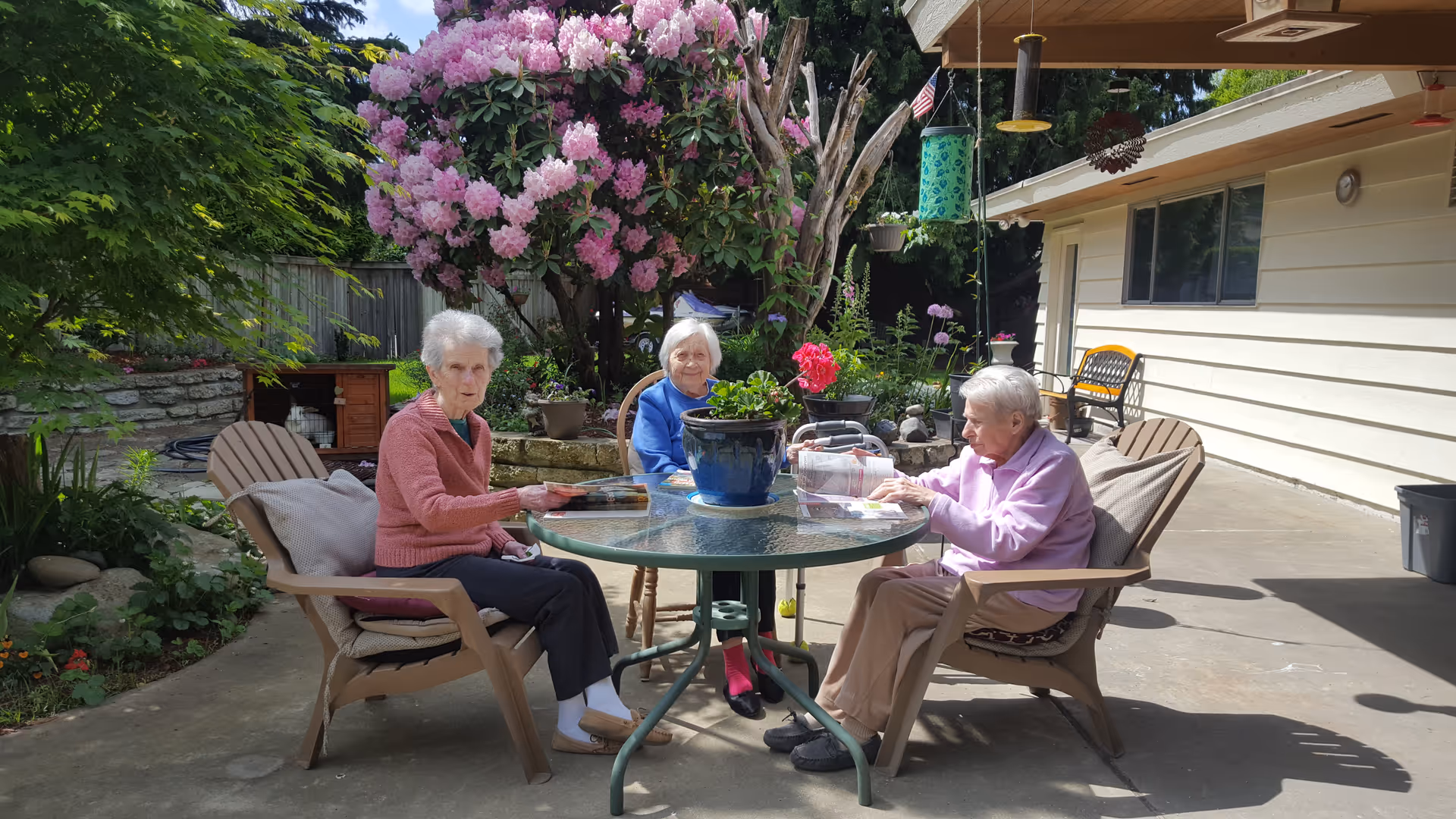 Three elderly women sitting around a glass outdoor table in a garden patio area. They are engaged in reading or looking at papers. The patio is surrounded by greenery, including a large blooming pink rhododendron bush and other plants. The setting is sunny and peaceful with a beige building wall in the background.
