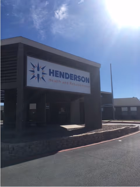 Exterior view of Henderson Health and Rehabilitation facility under a clear blue sky with the sun shining brightly. The building has a covered entrance with a large sign displaying the facility's name and logo.