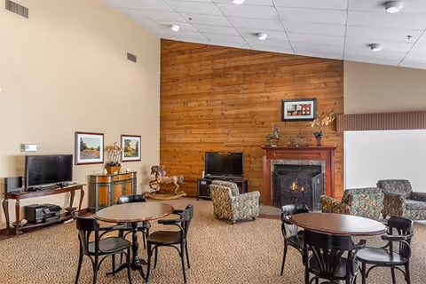 A cozy common area with a wooden accent wall featuring a fireplace and a flat-screen TV. The room has round tables with black chairs, patterned armchairs, framed artwork on the walls, and decorative items including a wooden horse sculpture and a vase with flowers.