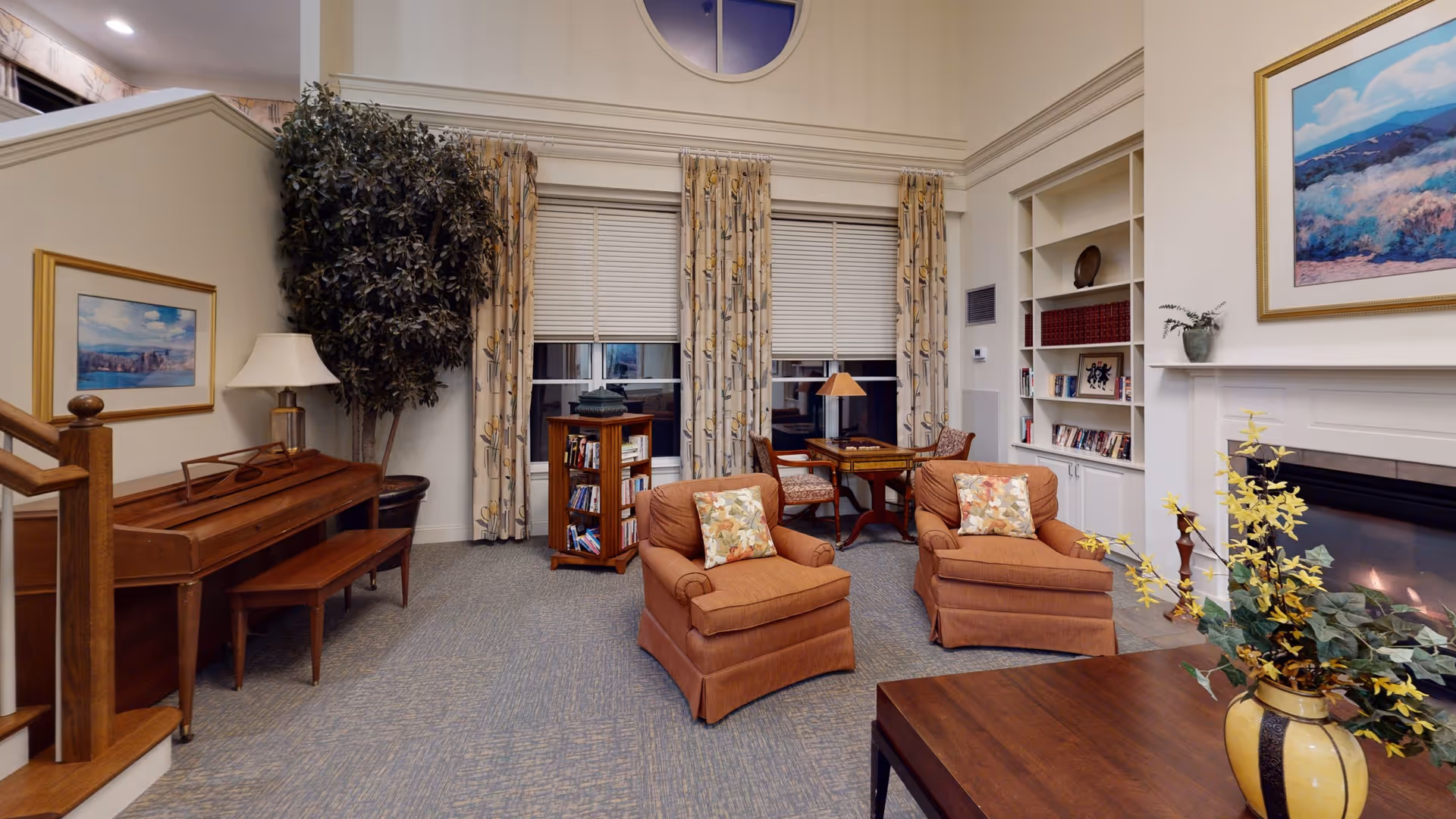 A cozy living room area with two orange armchairs featuring floral cushions, a wooden coffee table with a yellow vase and flowers, a fireplace with a painting above it, built-in white shelves with books and decor, a small table with two chairs near the window, a wooden piano with a bench, a large potted plant, and floral curtains covering the windows.