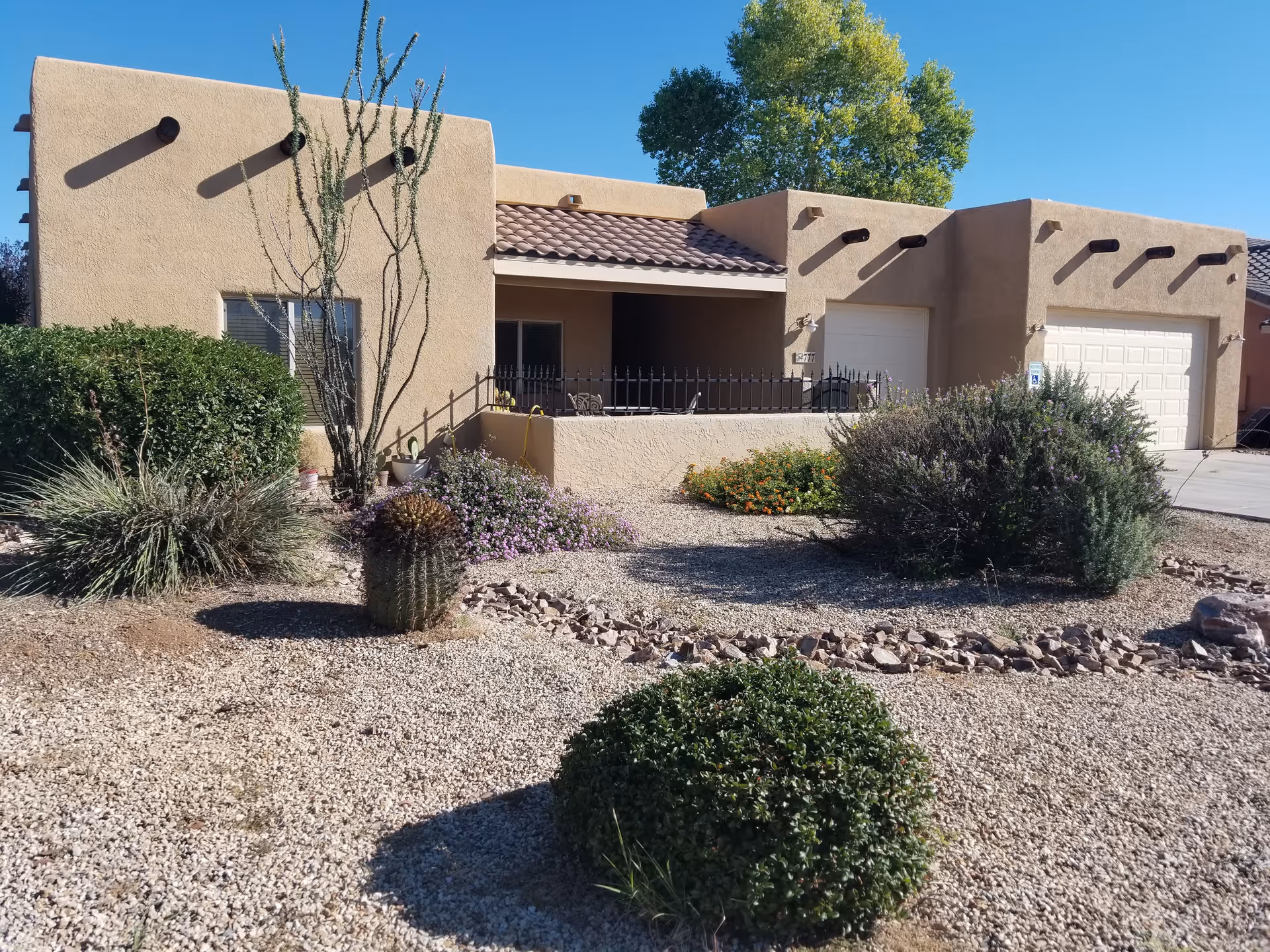 Exterior view of a single-story building with a beige stucco finish and a tiled roof. The front yard features desert landscaping with gravel, various bushes, cacti, and a small tree. There is a covered porch area with a black metal railing and two garage doors on the right side. The sky is clear and blue.