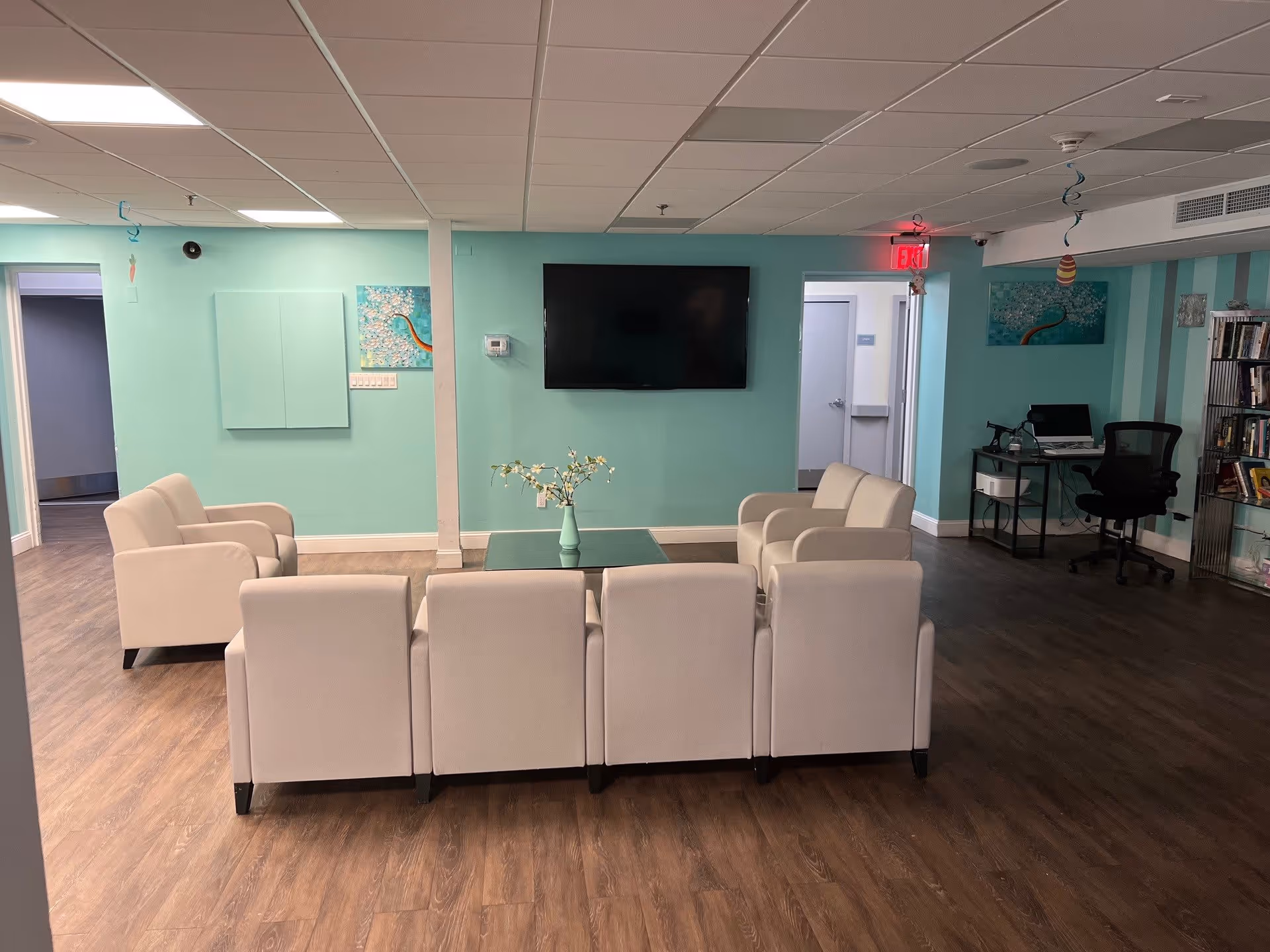 A cozy living room area with light beige armchairs arranged around a glass coffee table with a small vase of flowers. The walls are painted in a soft teal color, and a flat-screen TV is mounted on the wall. There is a desk with a computer and office chair in the corner, along with bookshelves filled with books. The floor is wood laminate, and there are two doorways visible leading to other rooms.