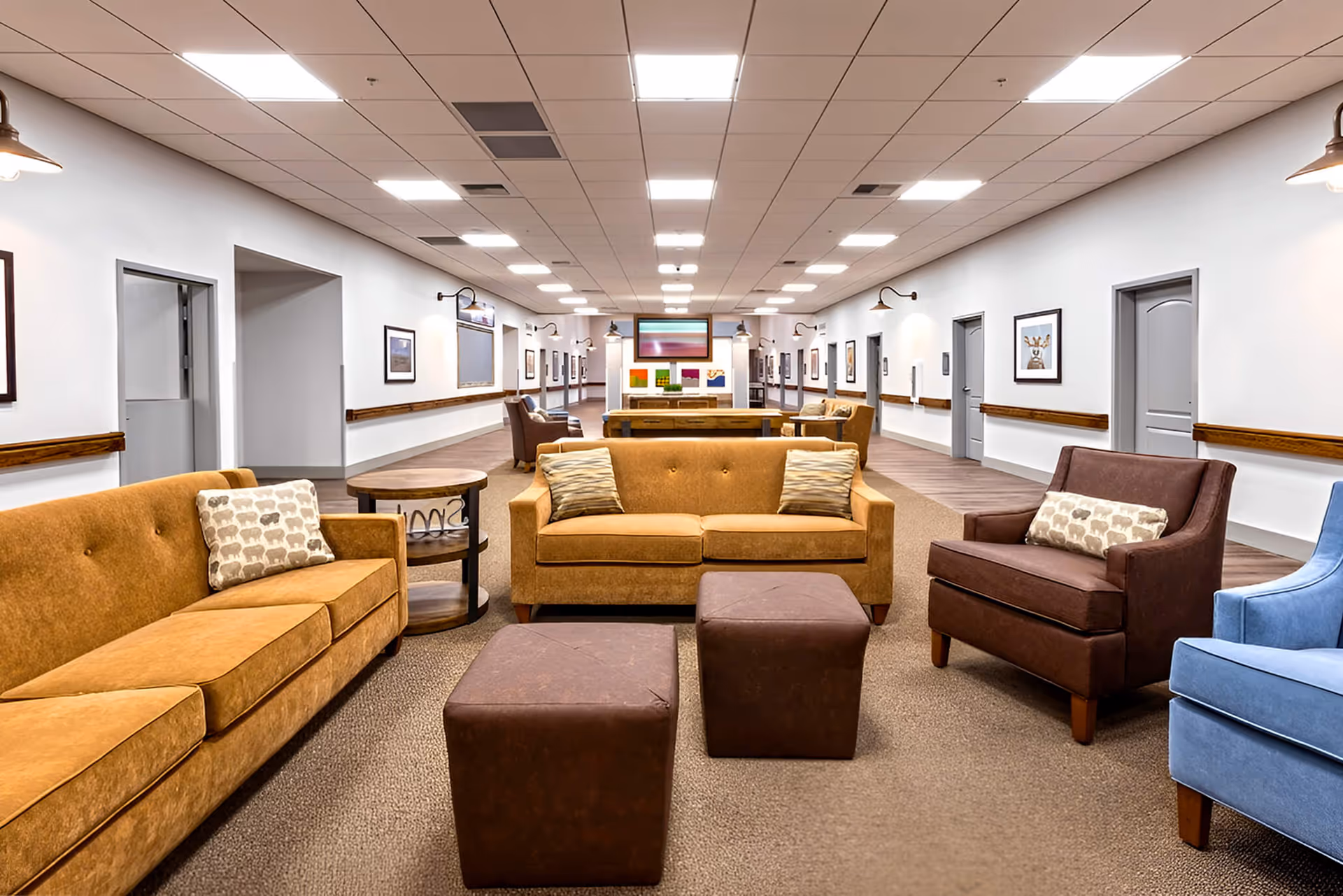 A spacious and well-lit common area in a senior living facility featuring comfortable seating including brown and blue armchairs, two brown sofas with patterned cushions, and two brown ottomans. The room has white walls adorned with framed artwork, wood handrails along the walls, and a long hallway with multiple doors leading to other rooms.