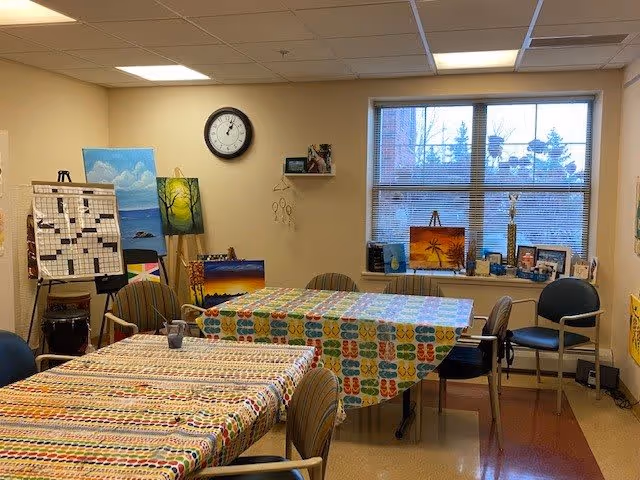 A cozy activity room with tables covered in colorful patterned tablecloths, surrounded by chairs. The walls display various paintings and artwork, including a large crossword puzzle board on an easel. A clock hangs on the wall above the artwork. A window with blinds lets in natural light, and several small decorative items are placed on the windowsill.