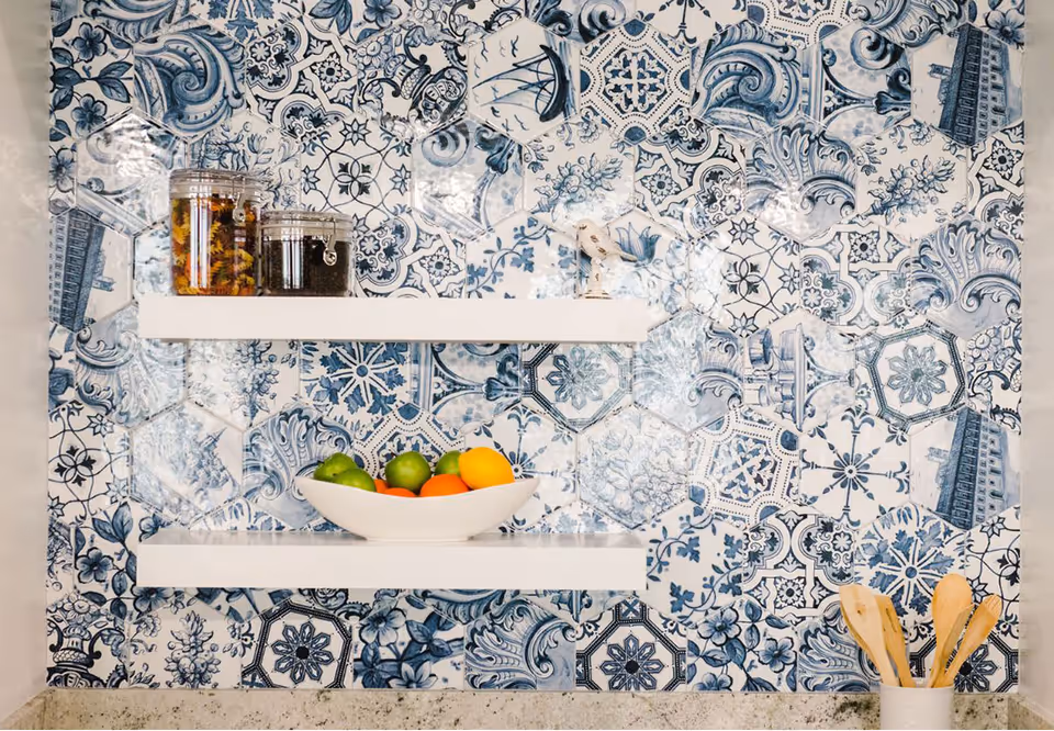 Two white floating shelves on a blue-and-white patterned tile backsplash holding a bowl of fruit, jars, and a container of wooden utensils.