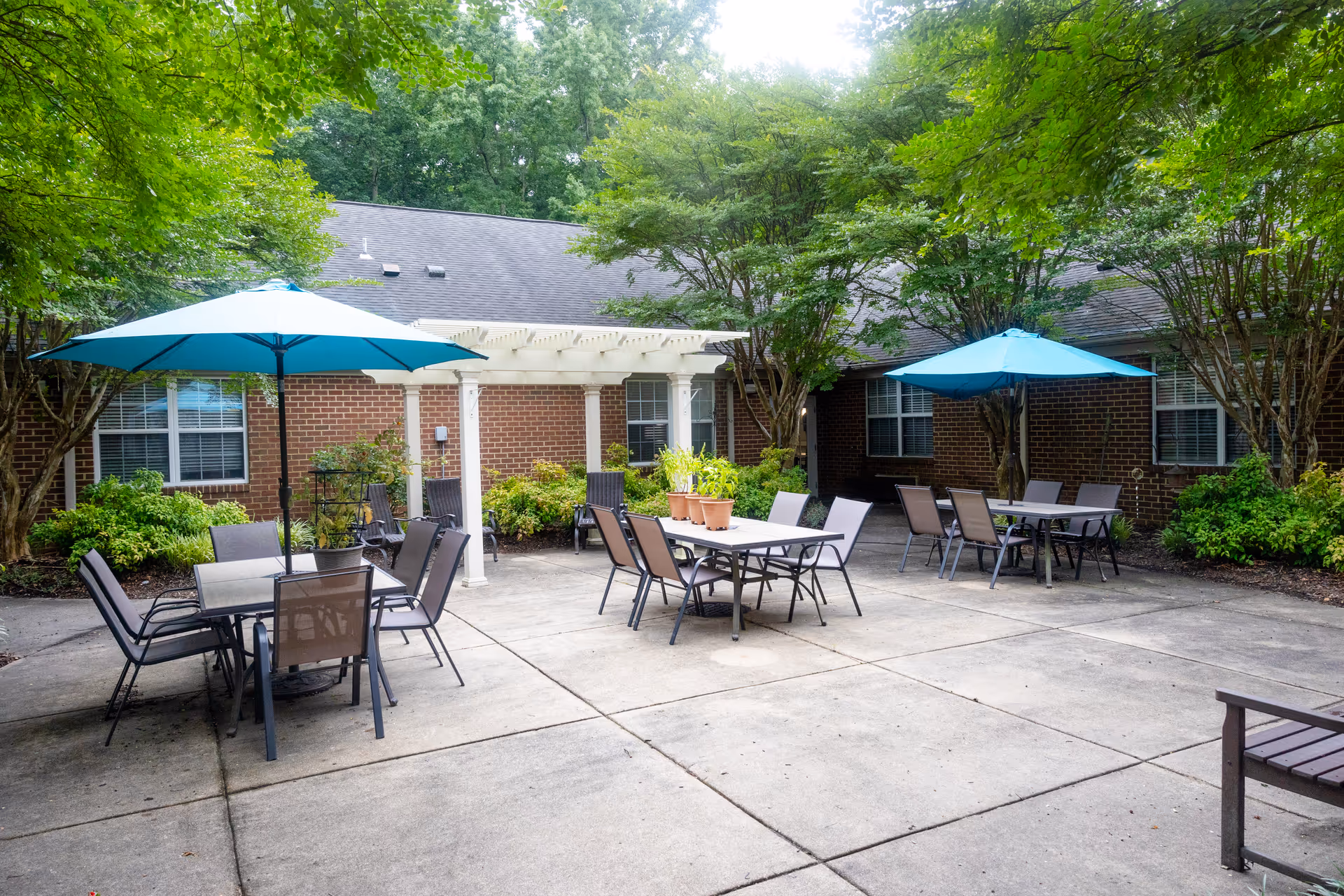 Outdoor patio area at Commonwealth Senior Living at Georgian Manor featuring multiple tables with chairs and blue umbrellas, surrounded by green trees and shrubs, with a brick building in the background.