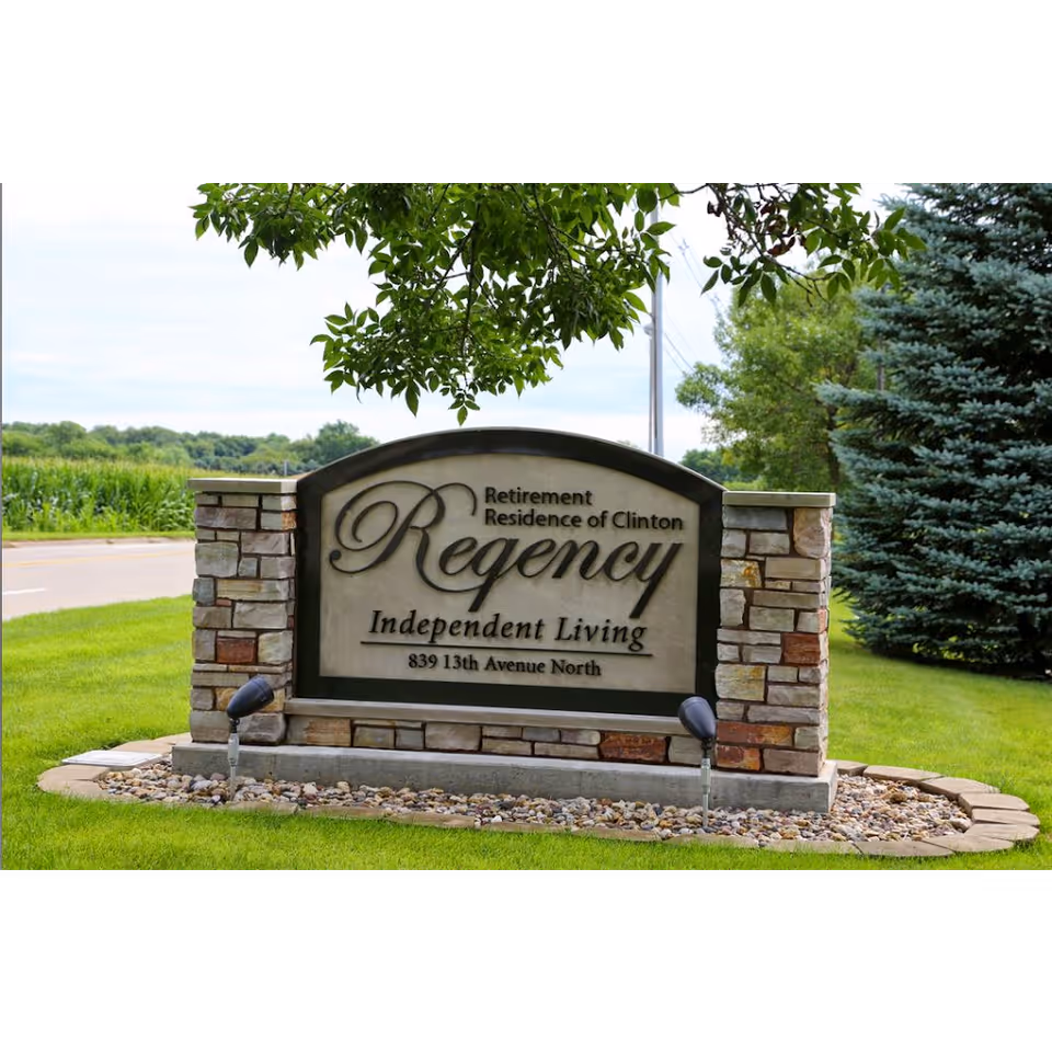 Stone and concrete sign for Regency Retirement Residence of Clinton Independent Living located on a grassy area near a road, with trees and a cornfield in the background.