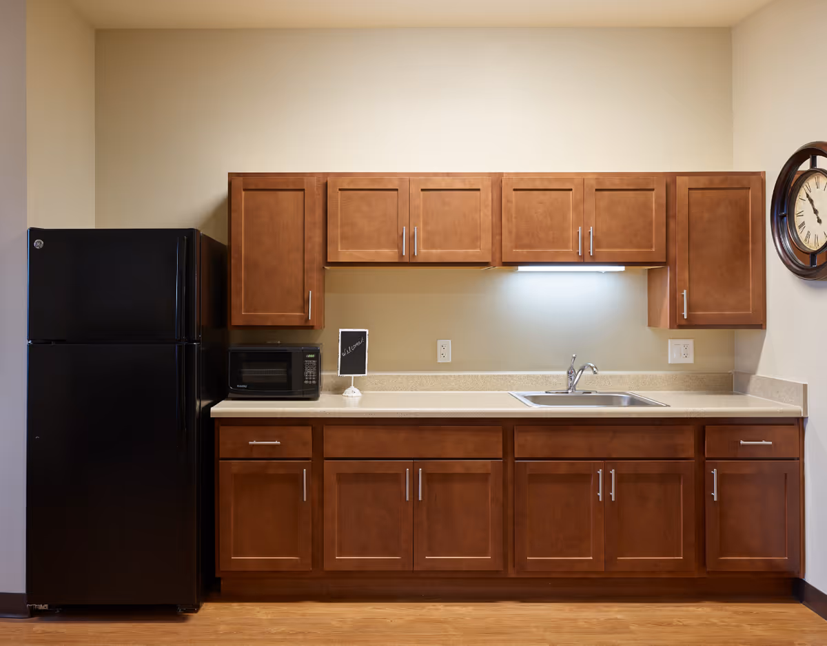A kitchen area with wooden cabinets, a black refrigerator on the left, a microwave on the countertop, a small sign that says 'Welcome', a sink with a faucet, and a wall clock on the right side.