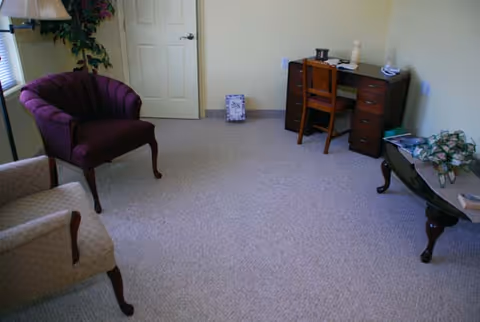 Carpeted sitting area with upholstered chairs, a small wooden desk, and a side table in a senior living facility room.