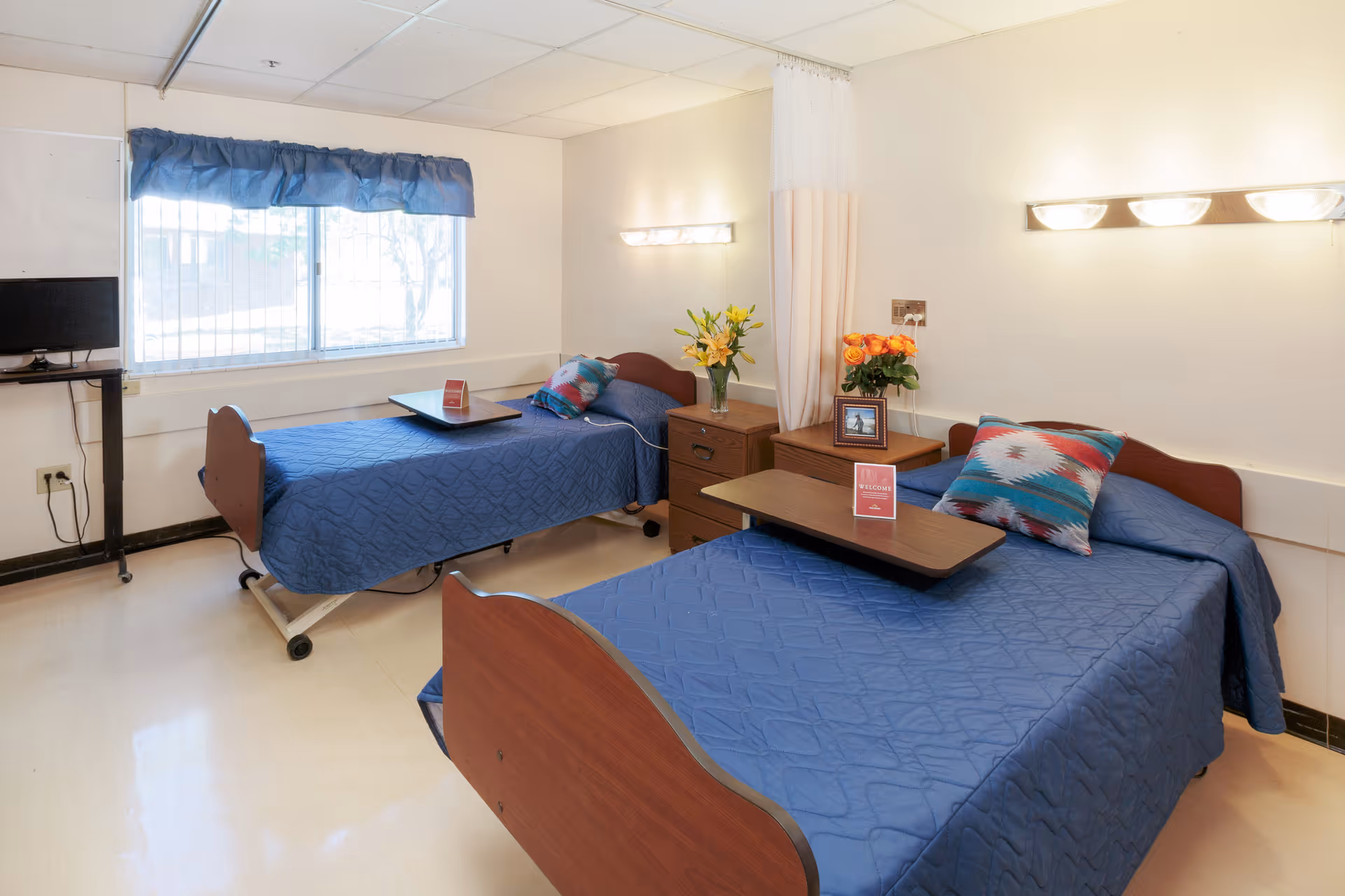A bright and clean room with two single beds covered in blue quilts, each with a colorful pillow and a wooden overbed table. Between the beds are two wooden nightstands with flowers and a framed photo. A window with blue curtains lets in natural light, and a small TV is mounted on the wall to the left.