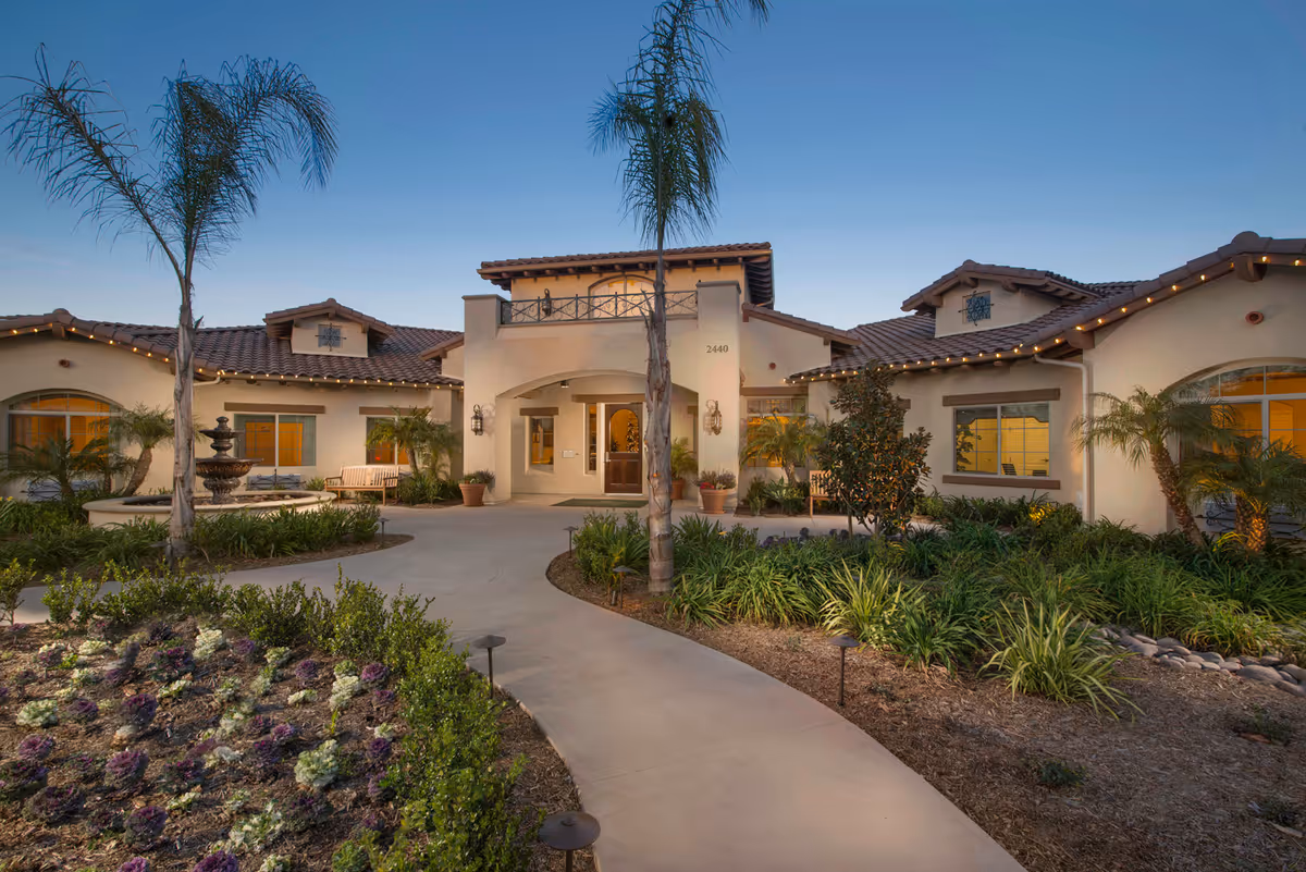 Exterior view of a single-story senior living facility building with a tiled roof, arched entrance, and well-maintained landscaping including palm trees, shrubs, and a flower bed. A paved walkway leads to the entrance, and warm lights illuminate the windows and building exterior at dusk.