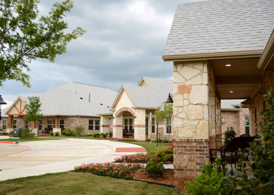 Exterior view of Mustang Creek Estates of Burleson showing a landscaped driveway with flower beds, trees, and a building with stone and brick facade under a cloudy sky.