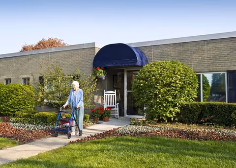 An elderly woman using a walker is walking on a sidewalk in front of a single-story brick building with a blue awning over the entrance. The building is surrounded by well-maintained bushes, flower beds, and green grass under a clear blue sky.