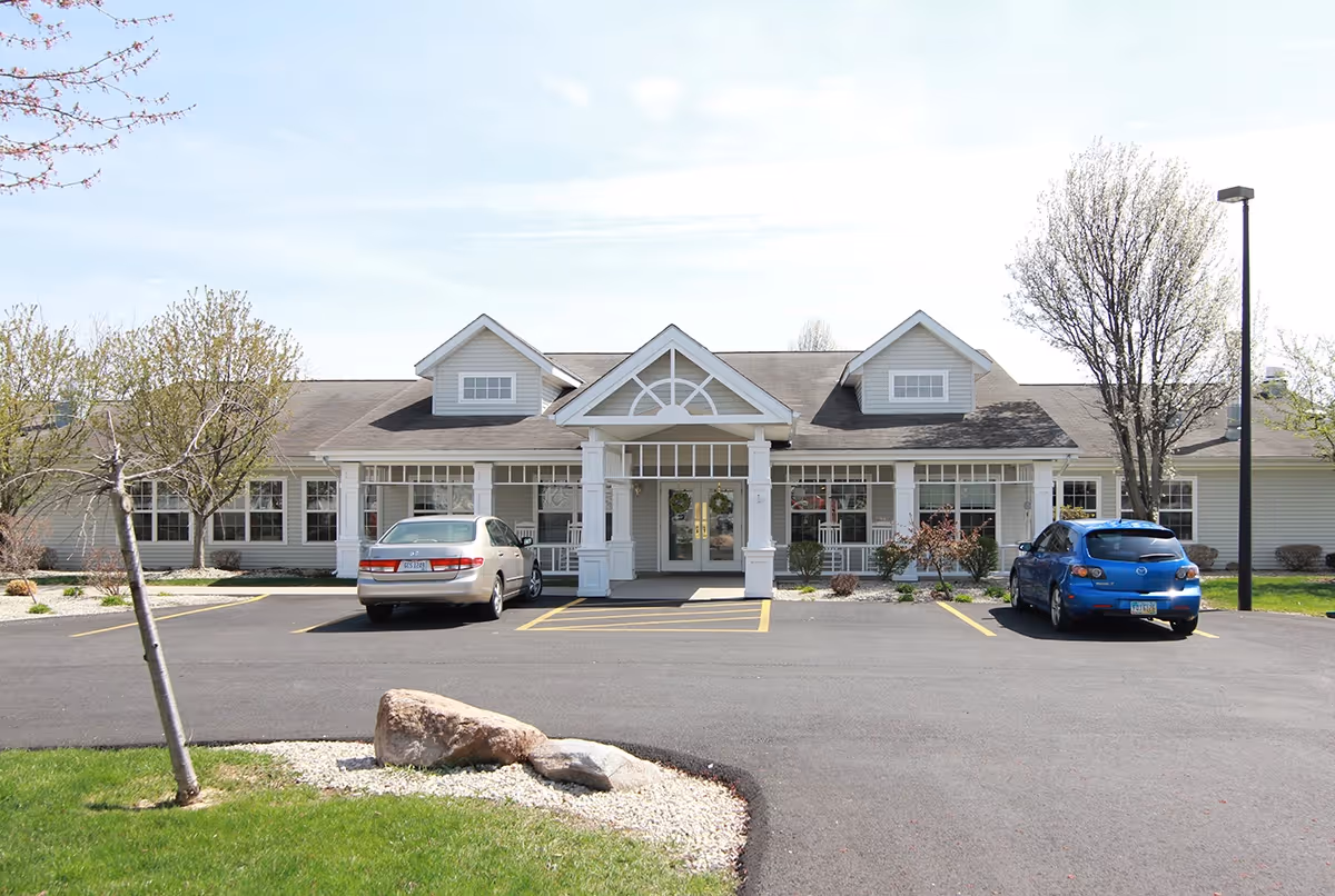 Front exterior view of a single-story building with a covered entrance supported by white columns, two cars parked in front, and trees and landscaping around the parking area under a clear sky.
