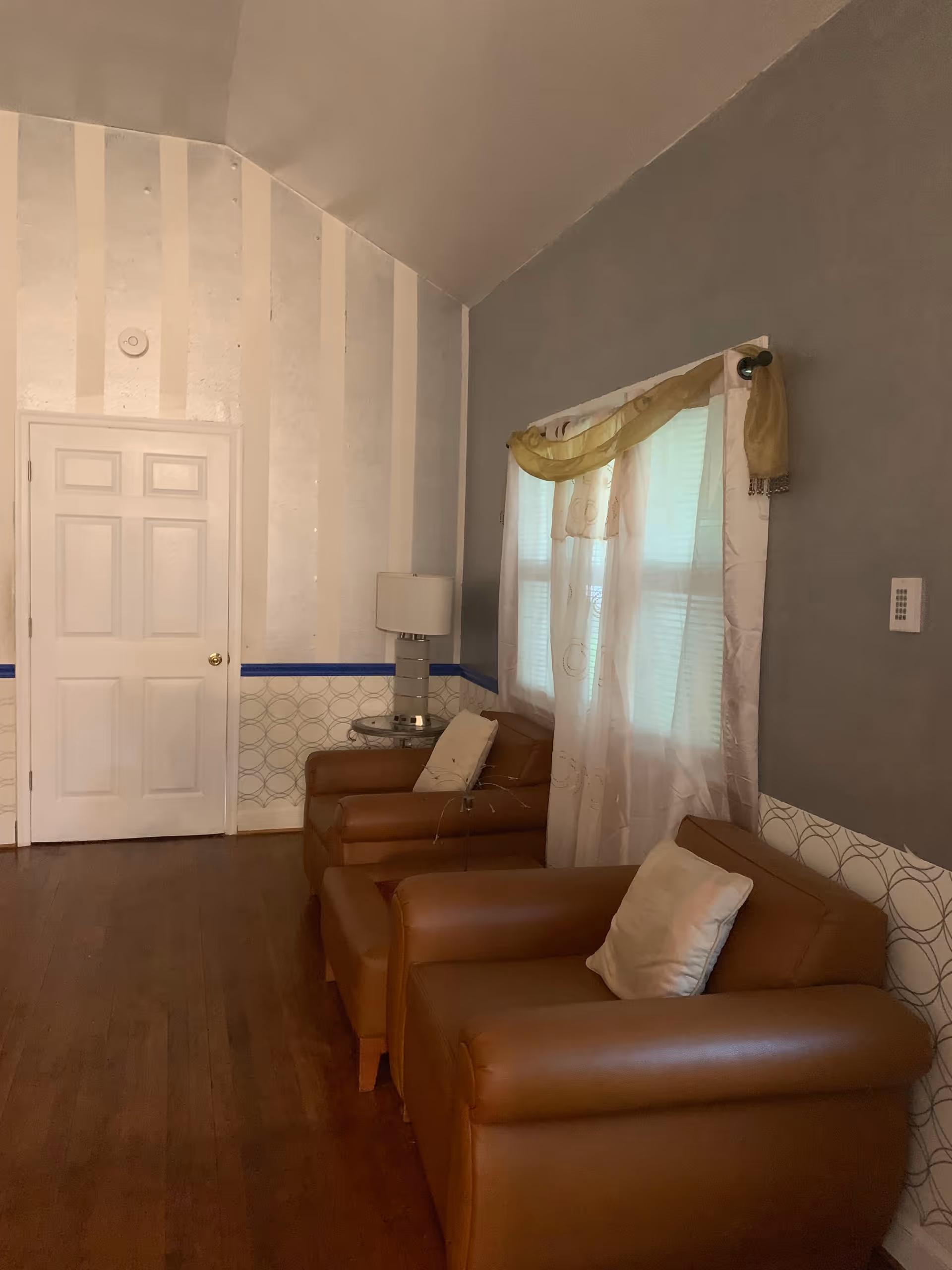 A cozy interior room with two brown leather armchairs each with a white pillow, a small round glass side table with a modern lamp, a window with sheer white curtains and a decorative gold swag valance, and a white door on the left side. The walls have a two-tone design with a gray upper section and a patterned lower section separated by a blue border. The floor is wooden.