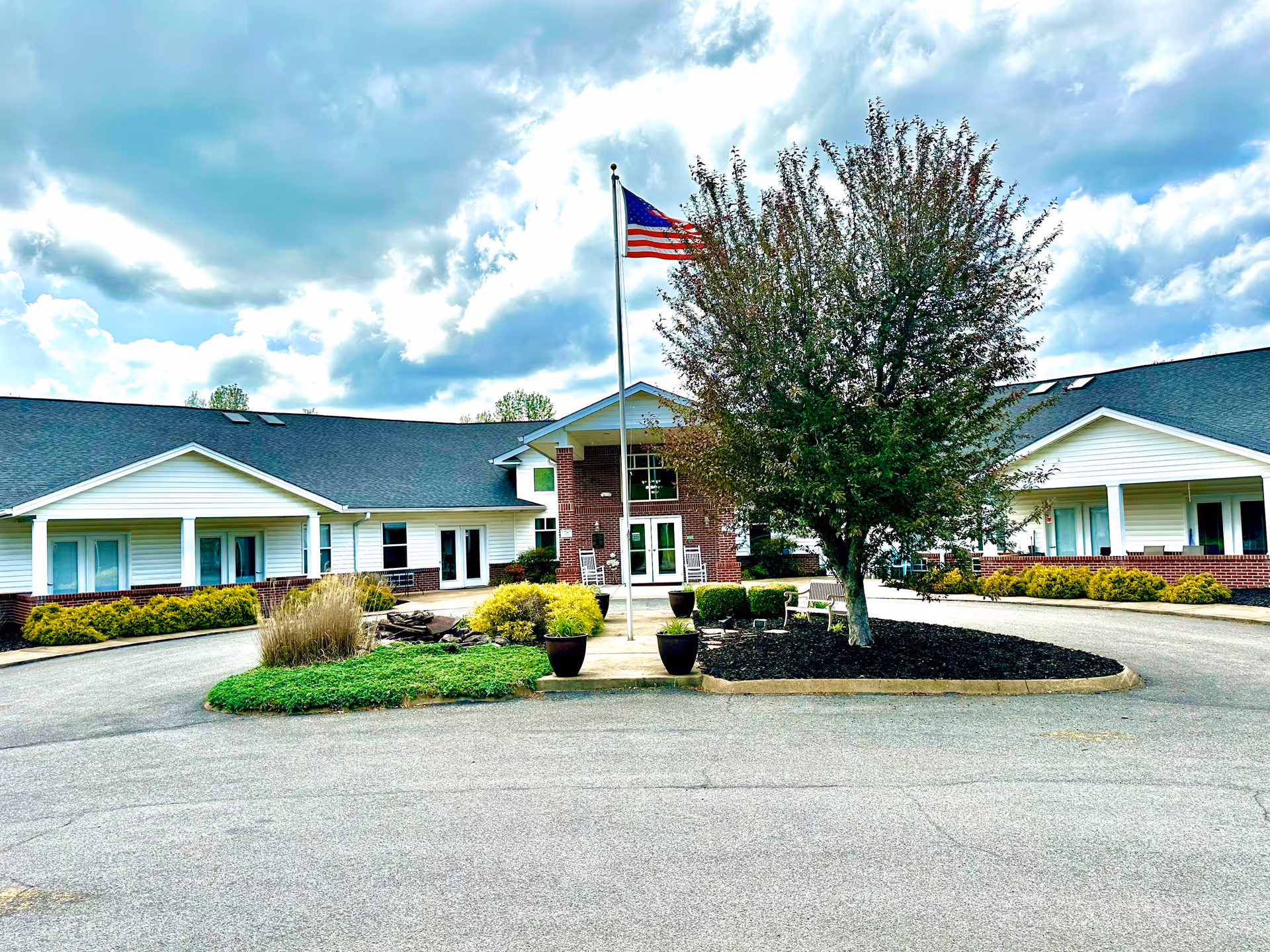 Front exterior view of Loving Care Assisted Living Facility showing a single-story building with white siding and brick accents. There is a circular driveway with a landscaped island in the center featuring a tree, shrubs, potted plants, and a flagpole flying the American flag. The sky is partly cloudy.