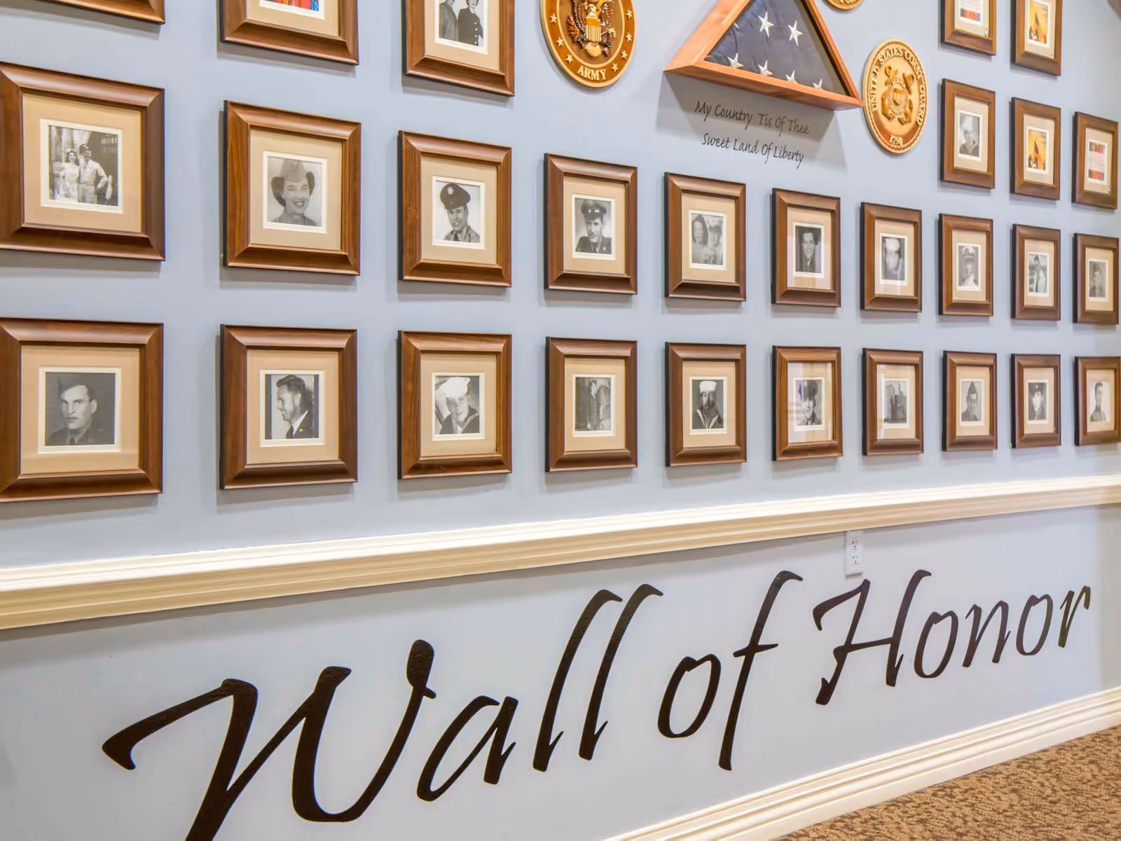 A wall display titled 'Wall of Honor' featuring multiple framed black and white photographs of military personnel, along with military insignias and a folded American flag in a triangular display case mounted on a light blue wall.