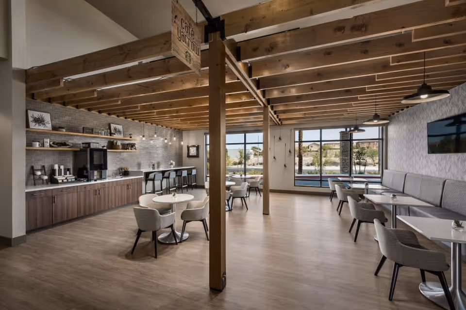 A spacious and modern dining area with wooden beams on the ceiling, light wood flooring, and large windows letting in natural light. The room features several tables with cushioned chairs, a long counter with bar stools, and a coffee station along one wall. The decor includes framed botanical prints and pendant lighting.