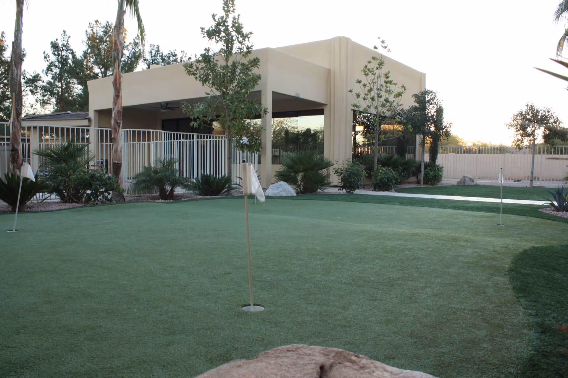 Outdoor putting green with three golf holes and flags, surrounded by palm trees, shrubs, and a beige building with large windows and a covered patio area in the background.