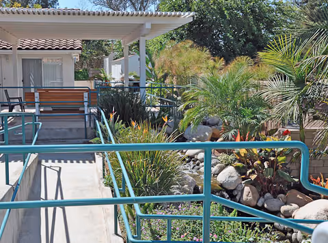 Outdoor garden area with a ramp and teal handrails leading to a covered porch attached to a building. The garden features various plants, rocks, and flowers, with trees and greenery in the background under a clear sky.