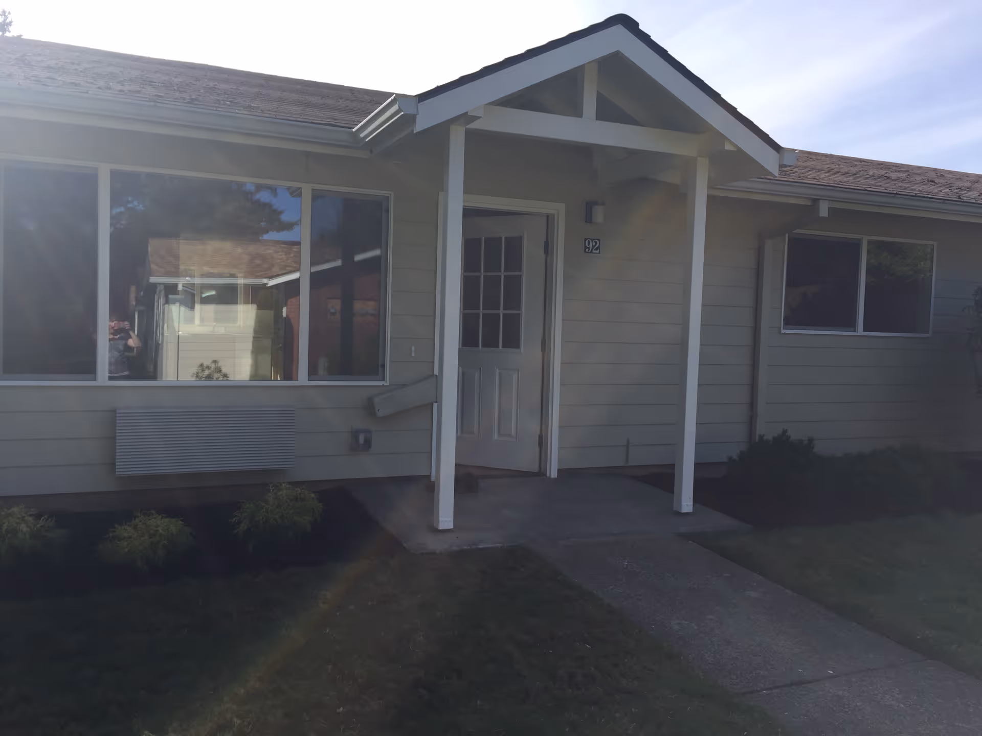 Exterior view of a single-story building with beige siding, a white door with glass panels, and two large windows. There is a small covered porch supported by two white columns, a concrete walkway leading to the entrance, and some bushes planted along the building. The building number 92 is visible next to the door.