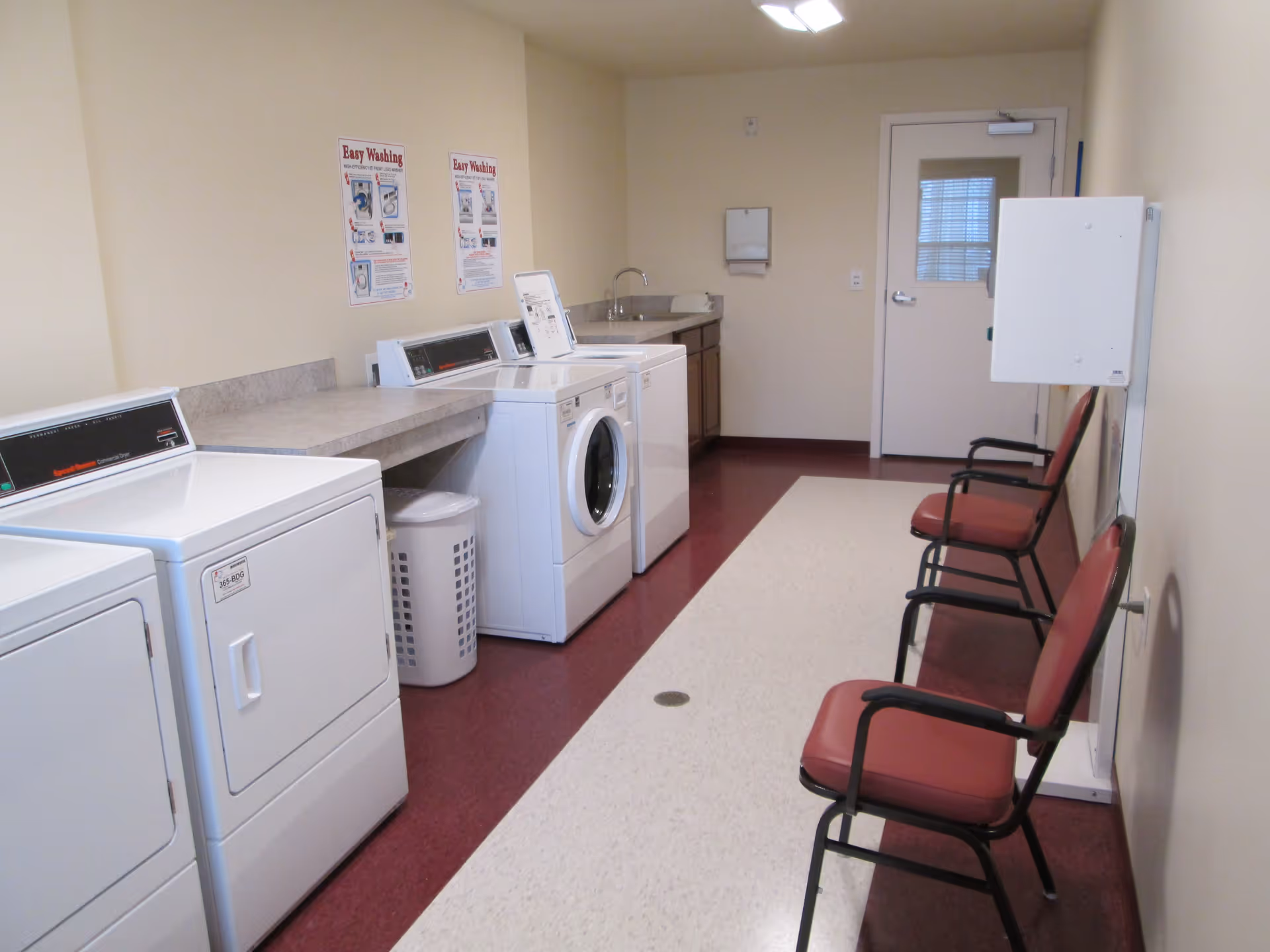 Laundry room with a washer, dryer, and laundry basket on the left side, a countertop with a sink at the back, and three red cushioned chairs along the right wall. The room has beige walls and a maroon and beige floor, with a door at the far end.