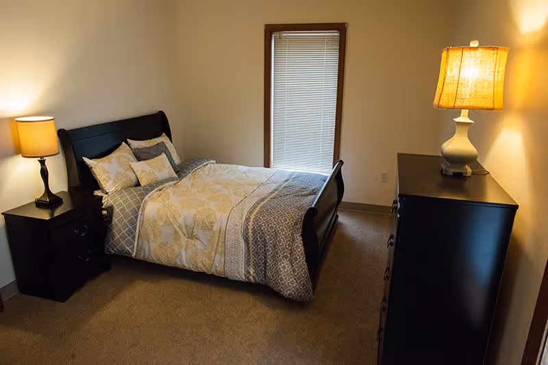 A bedroom with a neatly made bed featuring patterned bedding and multiple pillows. There is a wooden nightstand with a lamp on the left side of the bed and a tall dresser with a lamp on top on the right side of the room. A window with closed blinds is centered on the wall behind the bed.