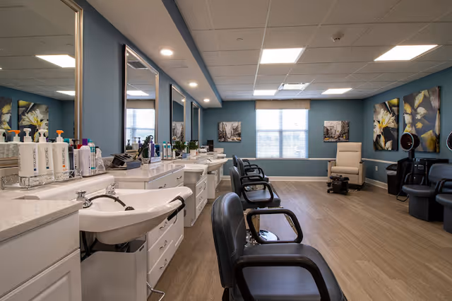 Interior view of a salon area in a senior living facility with multiple black salon chairs lined up along a counter with sinks and hair care products. The room has blue walls, large windows with blinds, framed artwork, and wood flooring.